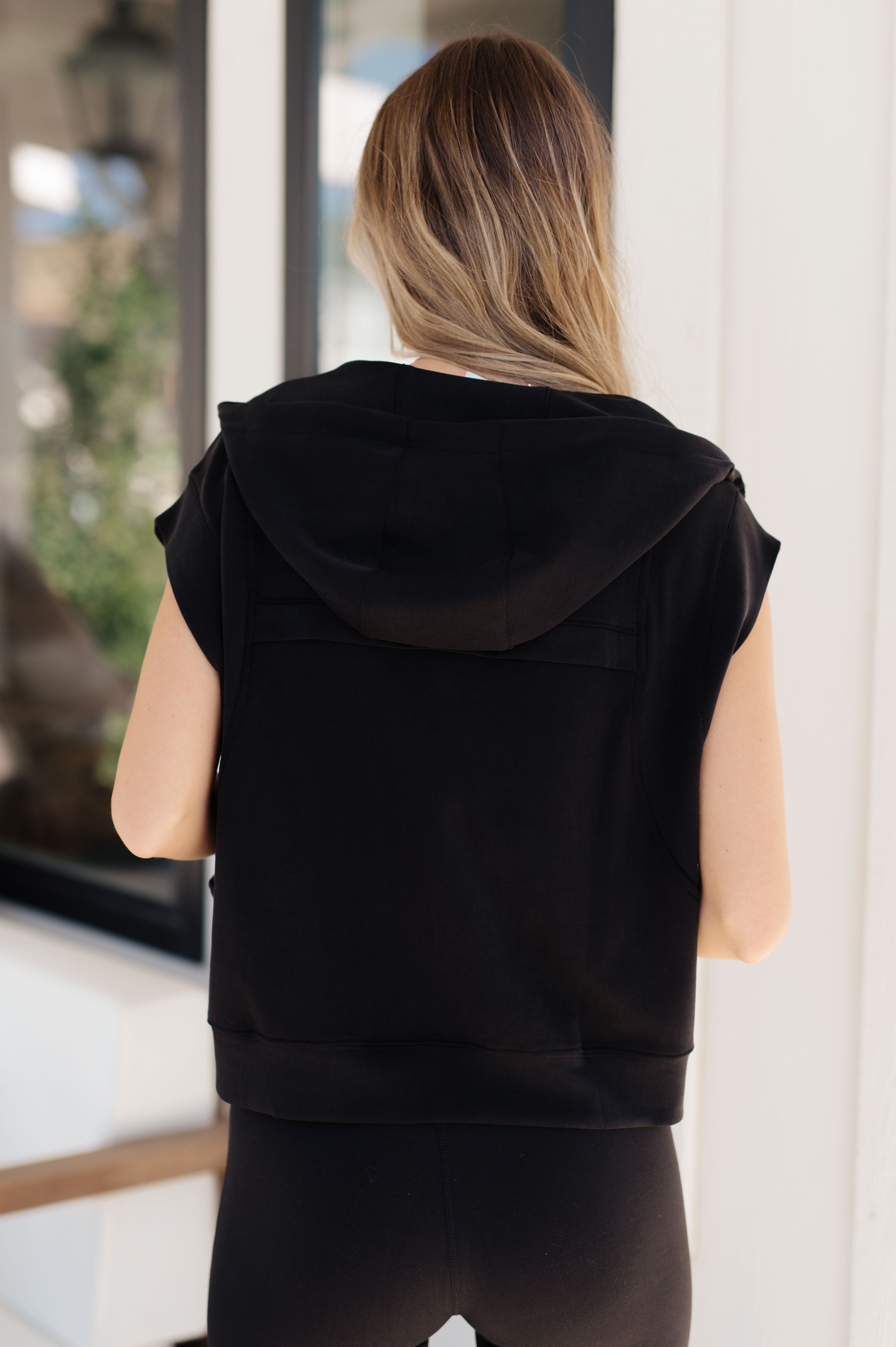 Woman with light brown hair wearing a black sleeveless hoodie seen from behind in a bright indoor setting