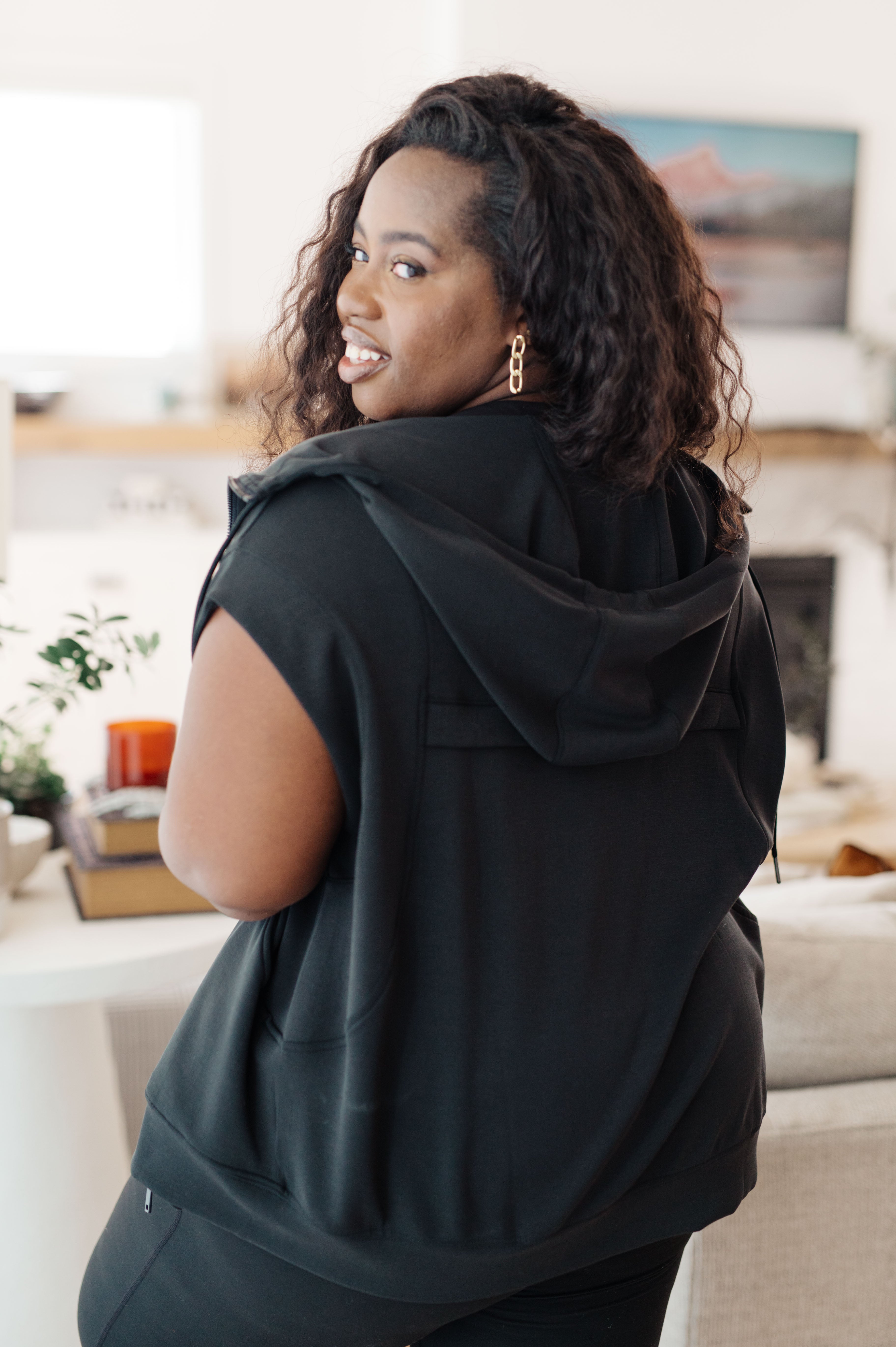 Curvy woman wearing a black sleeveless hoodie with a hood indoors, smiling over shoulder, accessorized with gold earrings