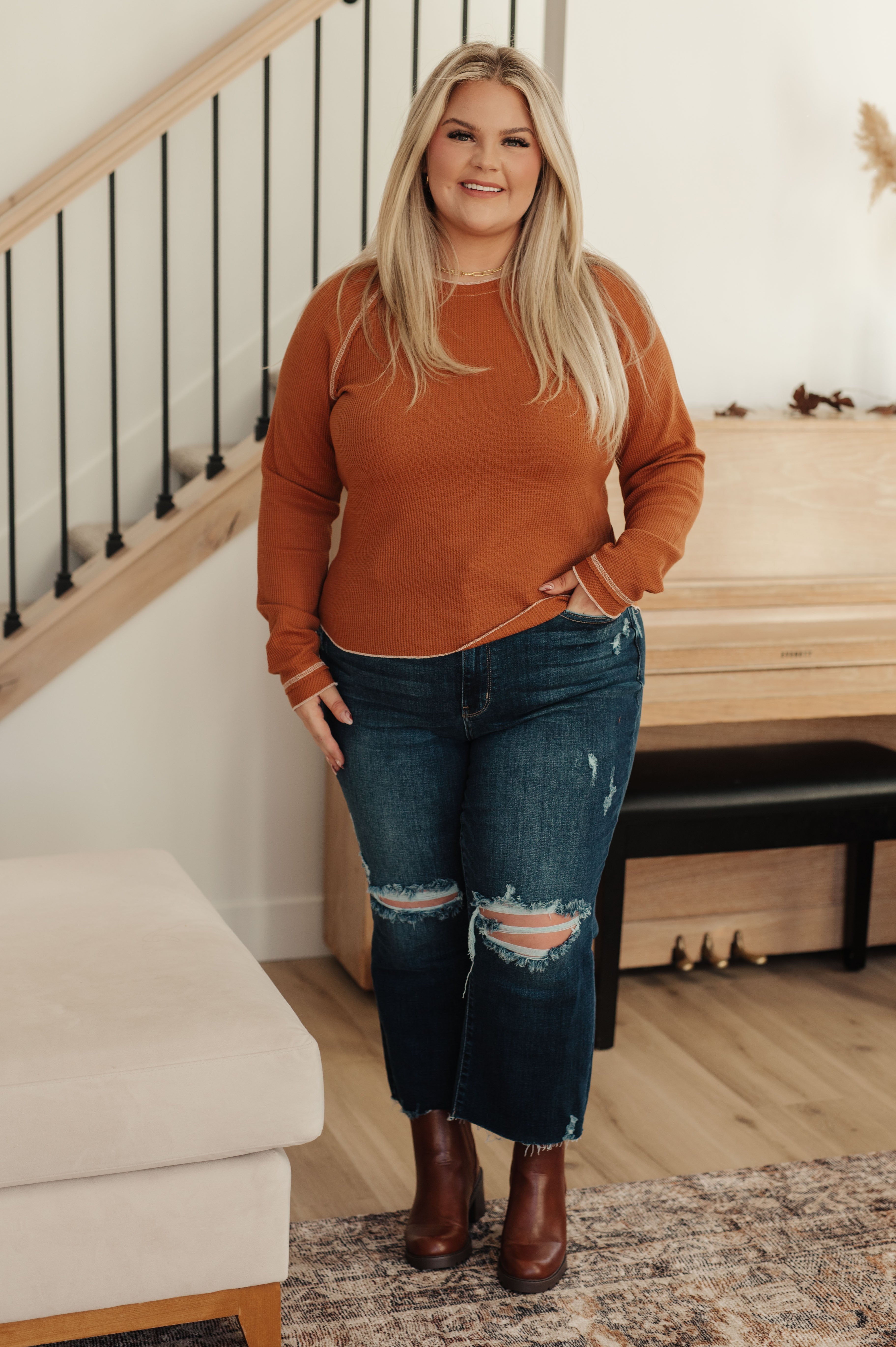 Plus-size woman wearing rust waffle knit top and distressed cropped jeans with brown ankle boots indoors, smiling