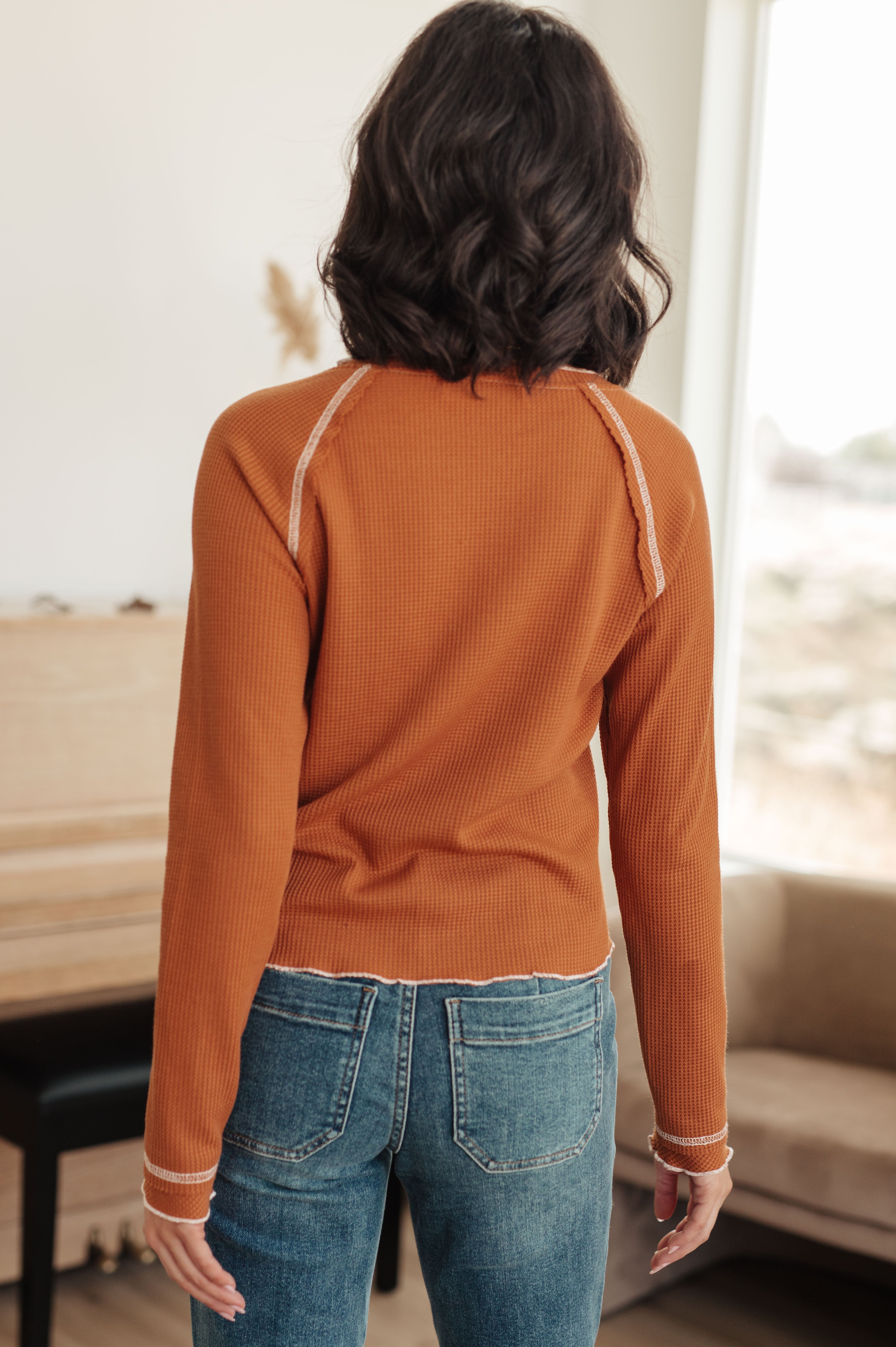 Woman wearing rust-colored waffle knit top with contrast stitching and blue jeans standing indoors by window