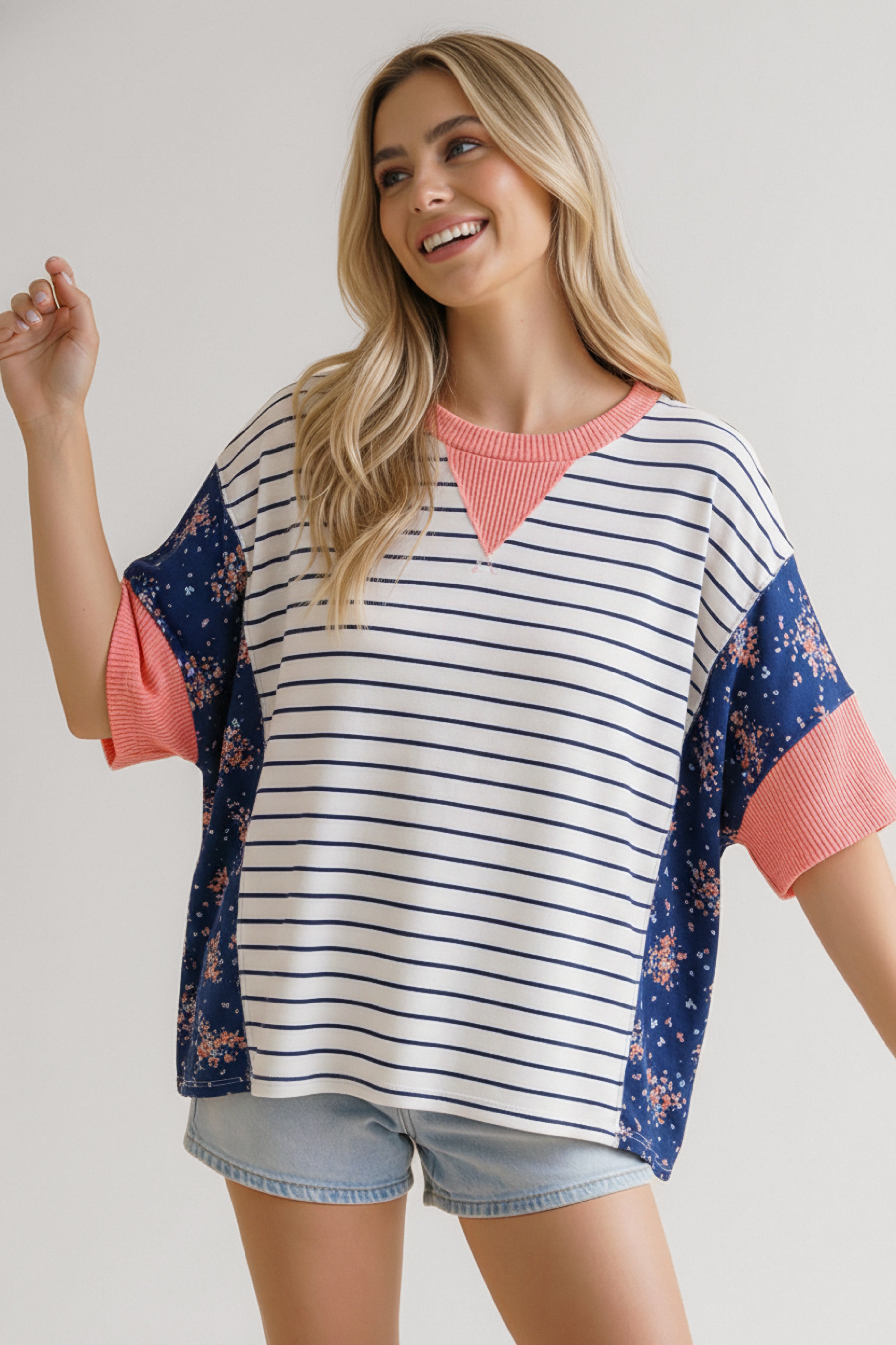 Smiling woman wearing blue striped top with floral side panels and coral ribbed neckline and sleeves paired with denim shorts
