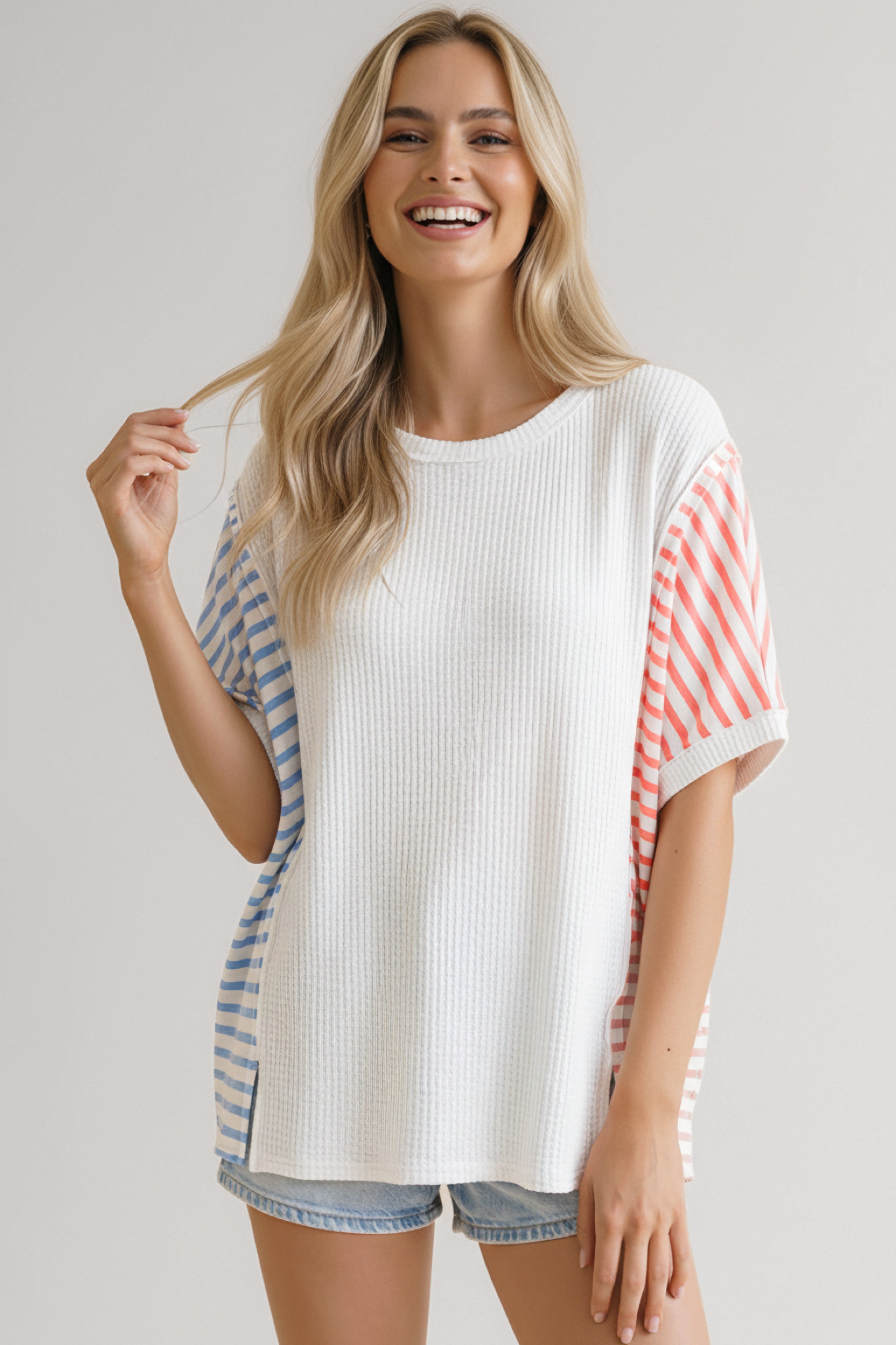 Smiling woman wearing ivory waffle knit top with blue and coral striped sleeves paired with denim shorts