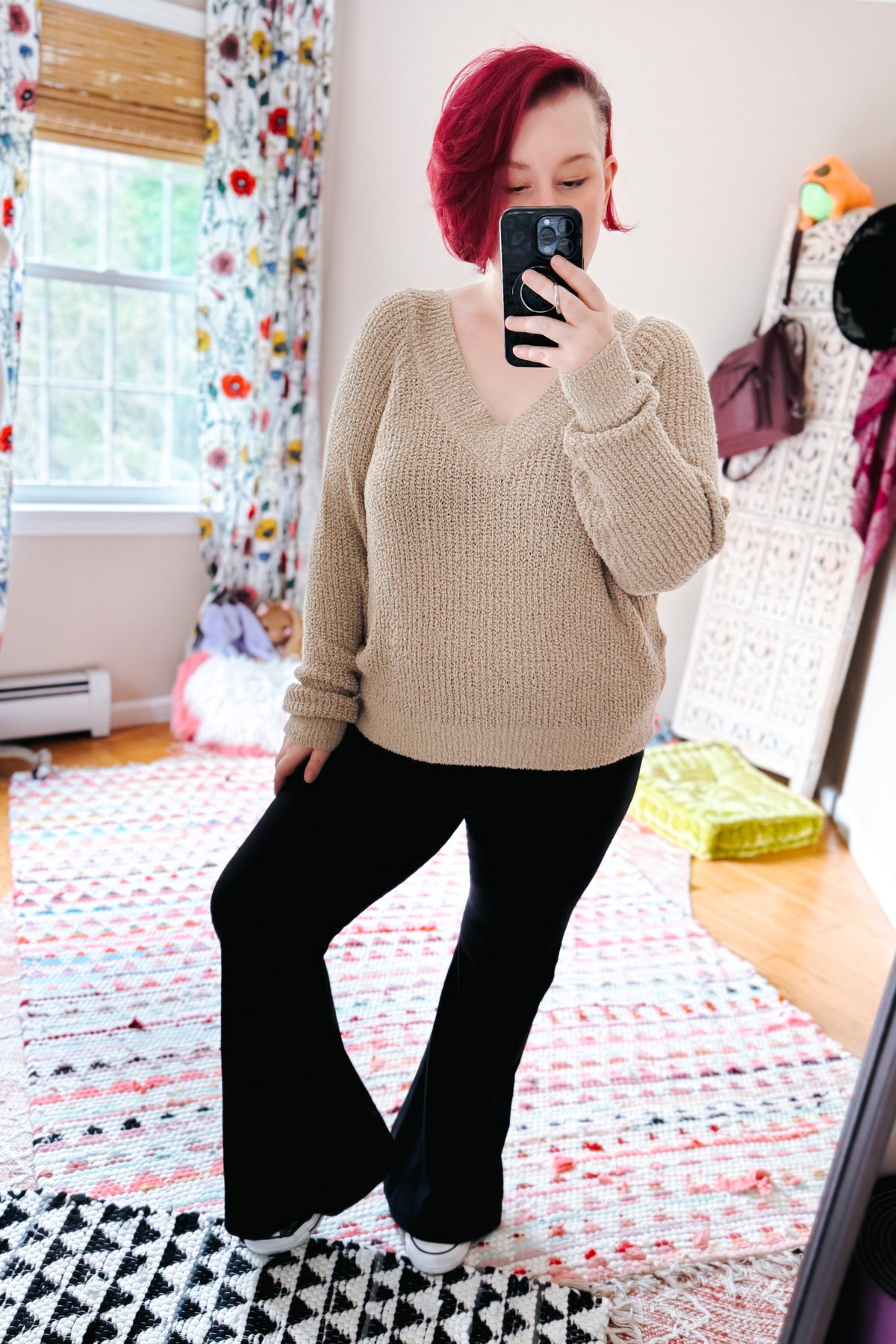 Red haired woman posing in a bedroom mirror selfie wearing a beige knit V neck sweater black flared pants and white sneakers