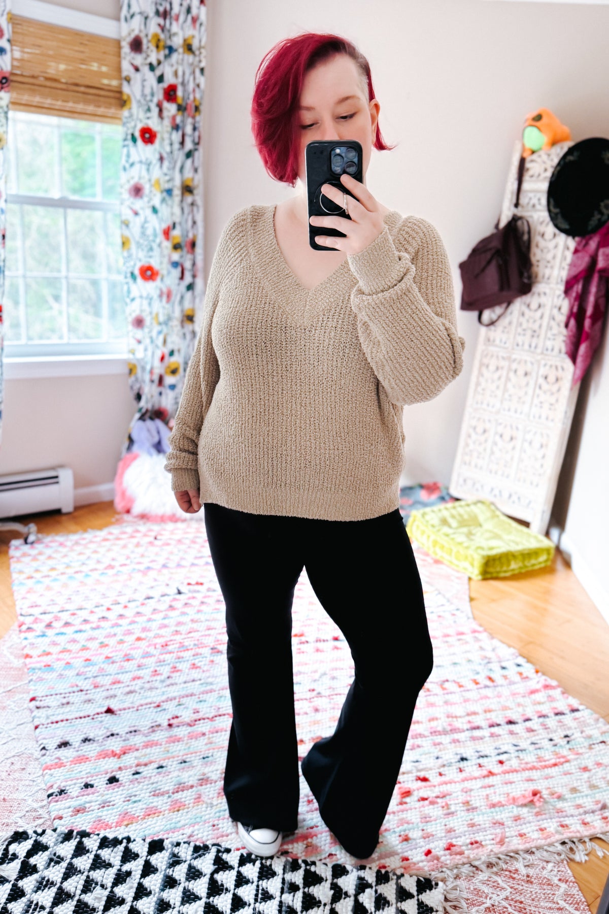 Red haired woman posing in a beige V-neck sweater with black flared pants and white sneakers during a bedroom mirror selfie