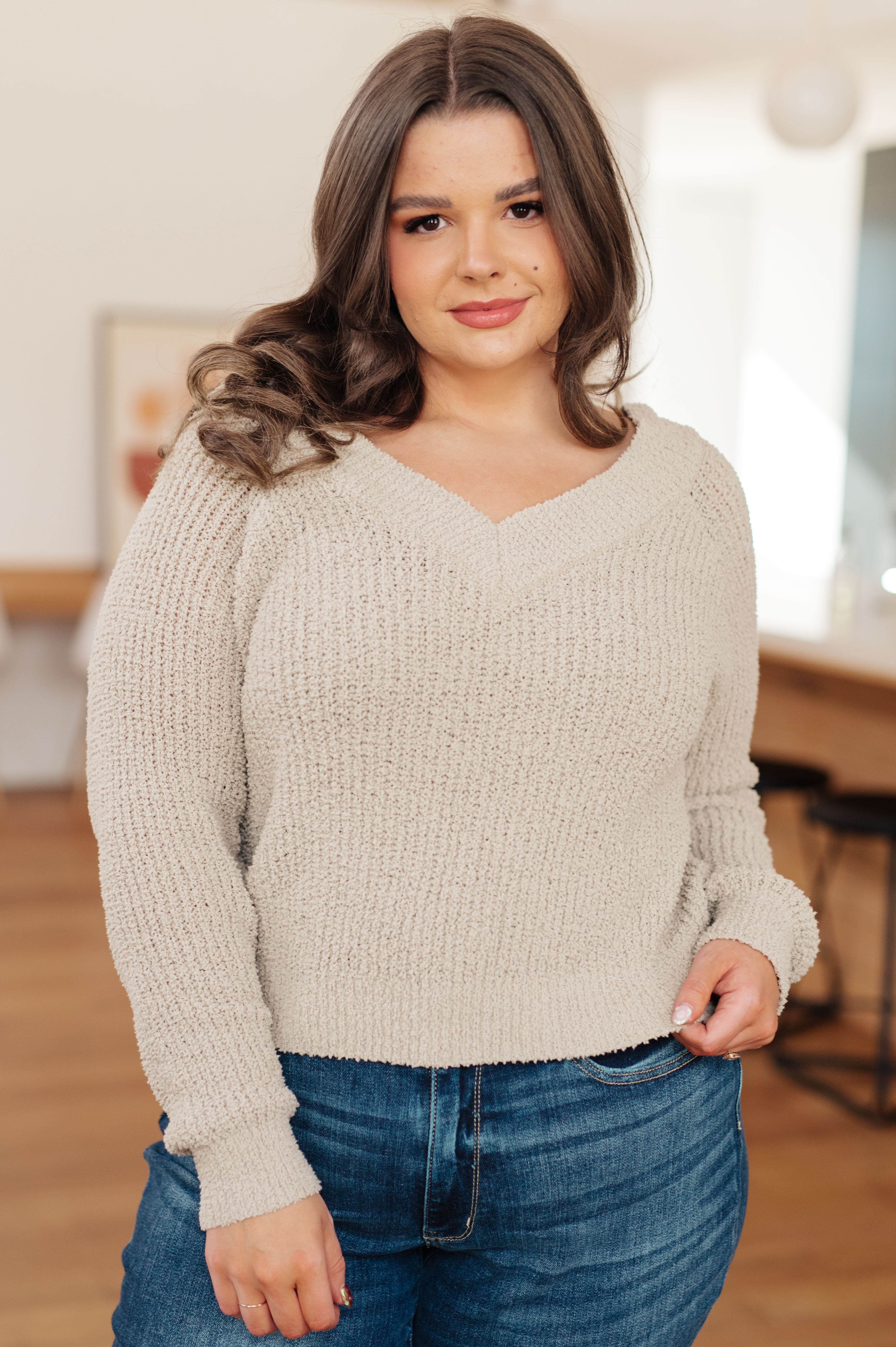 Woman wearing beige textured V-neck sweater with long sleeves and casual blue jeans in cozy indoor setting