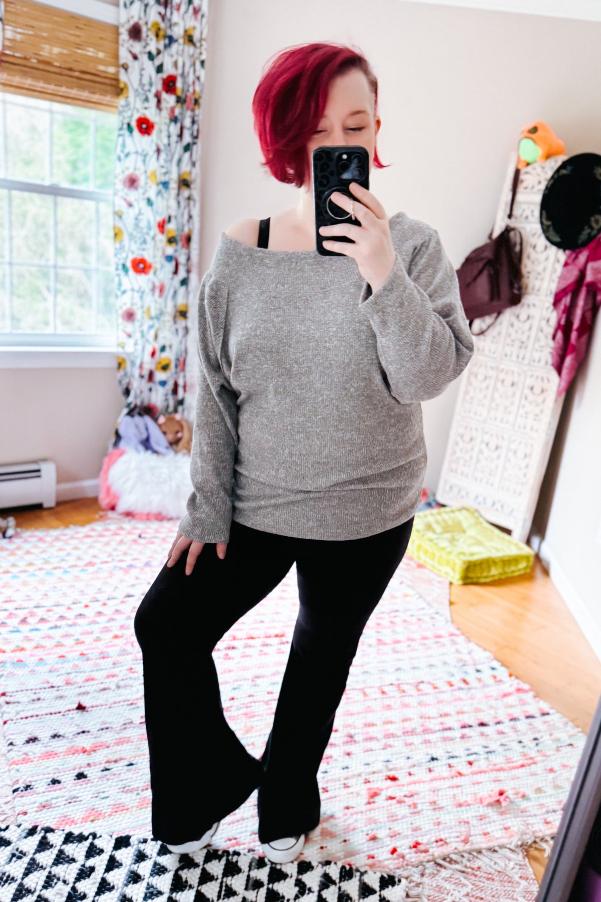 Woman taking a mirror selfie in a grey textured knit boat neck top with dolman sleeves worn off one shoulder and black pants indoors
