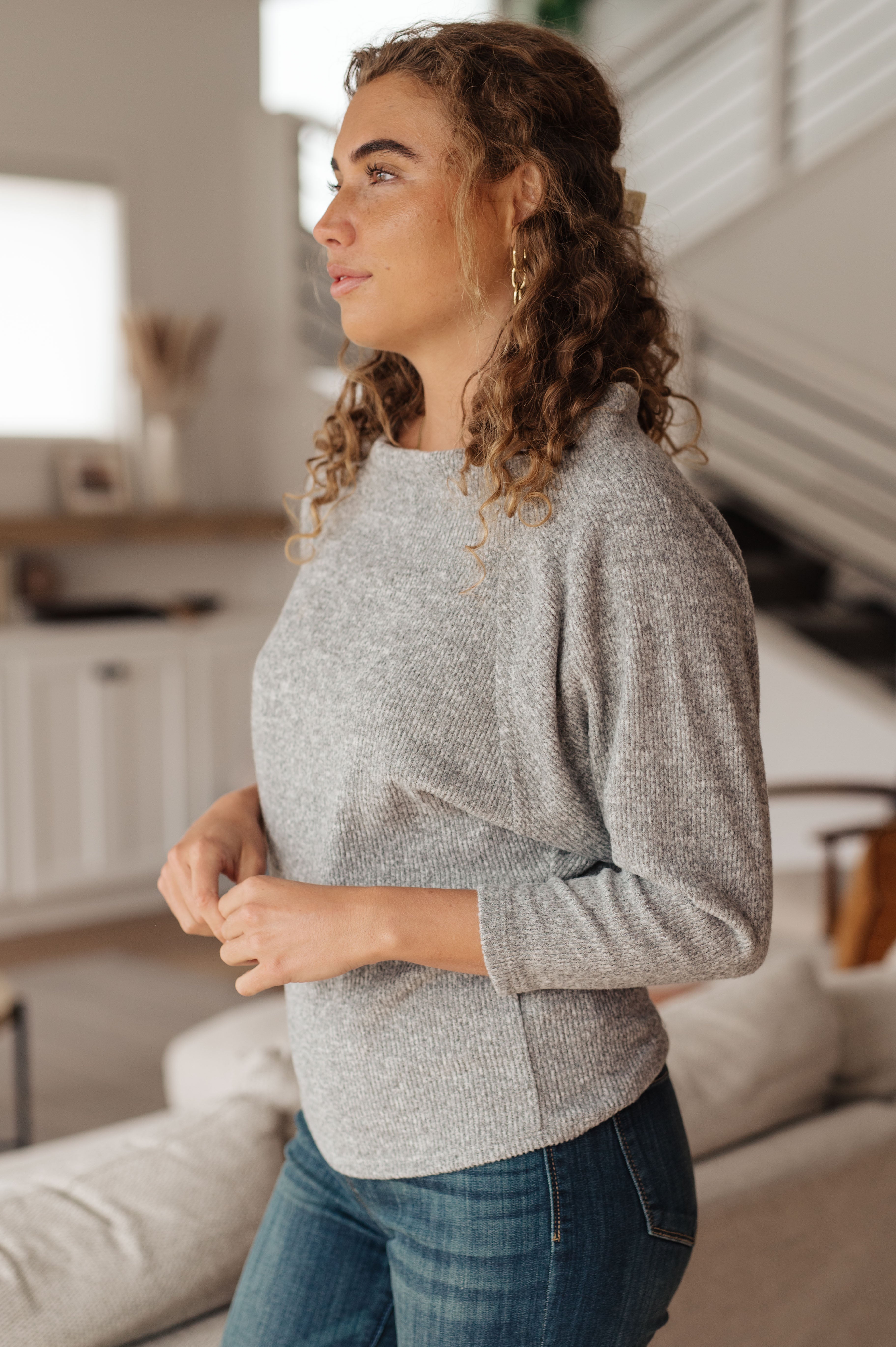 Woman with curly hair wearing a grey knit long sleeve top and blue jeans standing in a modern cozy room