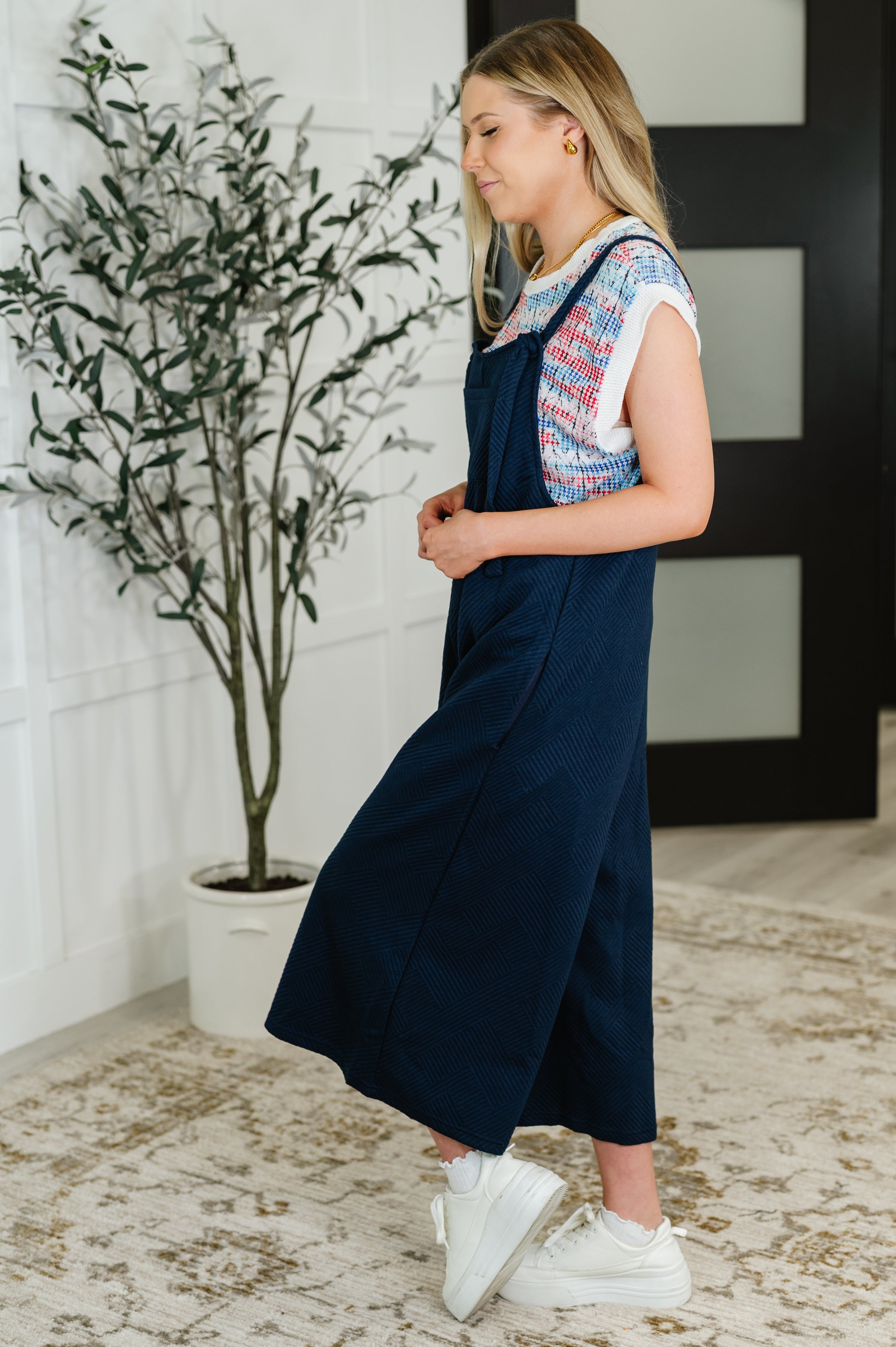 Woman wearing navy adjustable strap jumpsuit layered over a colorful knit top with white sneakers indoors