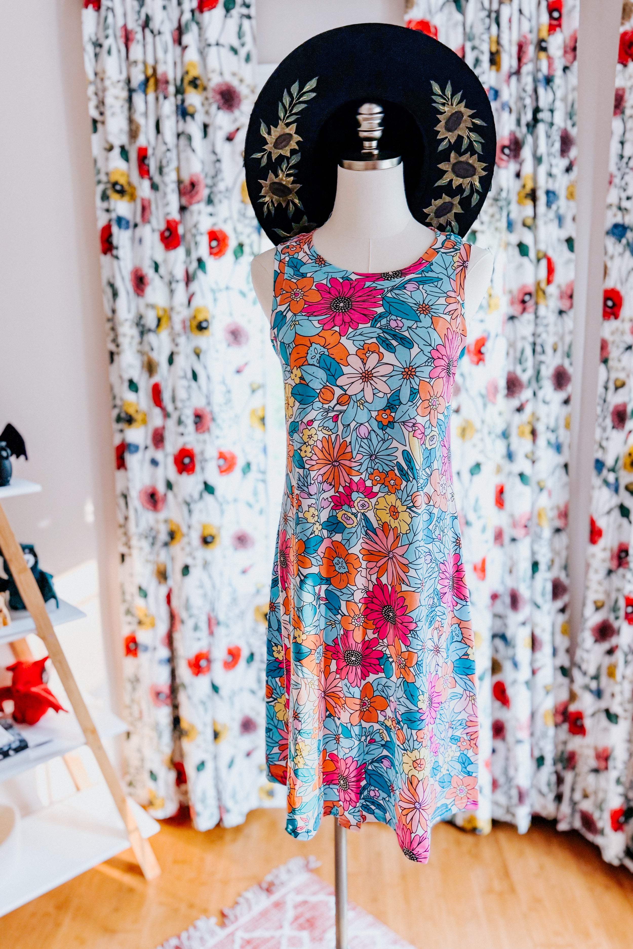 Mannequin wearing a colorful sleeveless floral swing dress with a wide-brimmed black hat decorated with sunflowers indoors