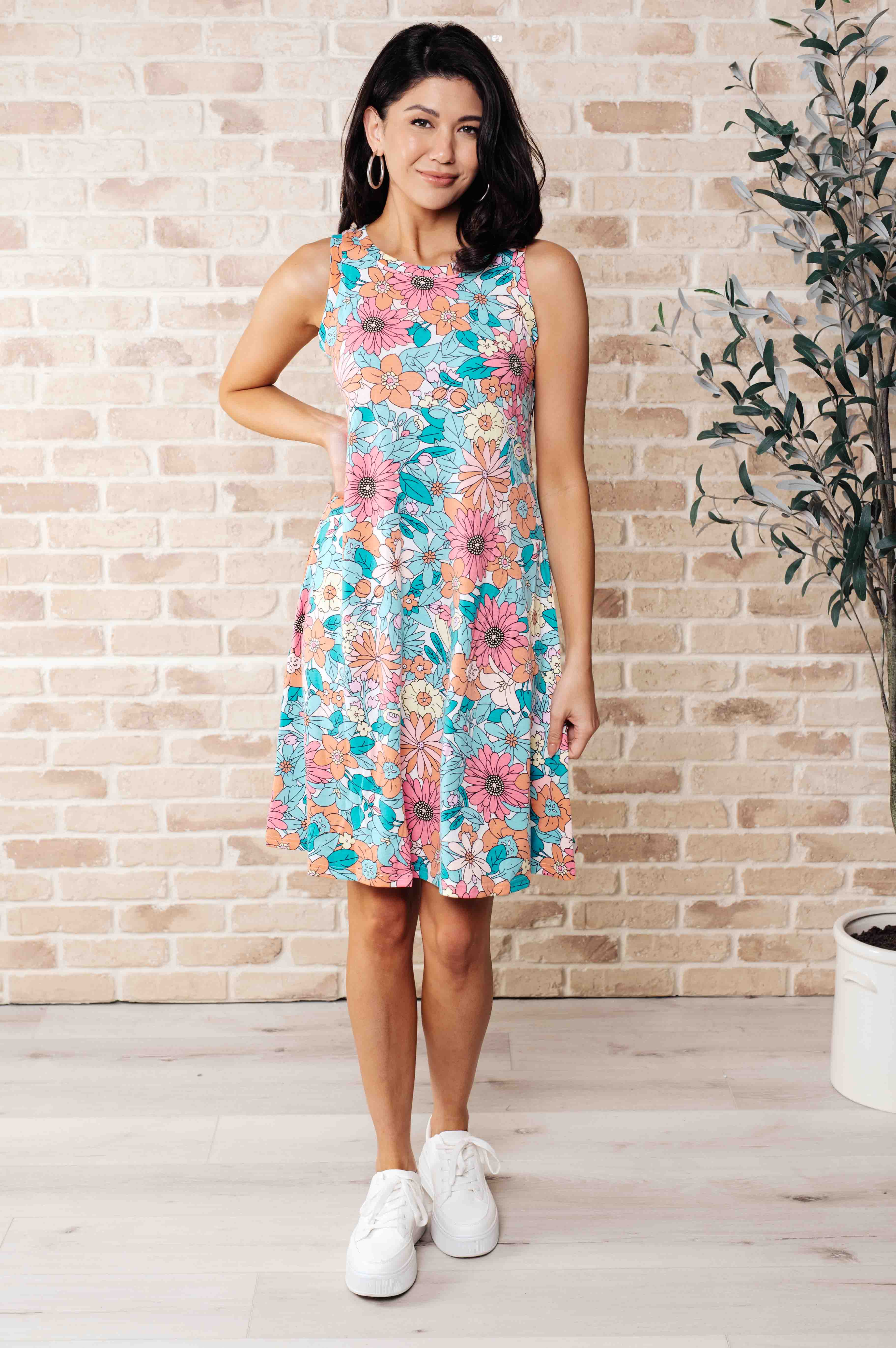 Woman wearing a sleeveless floral swing dress with blue, pink, and orange hues, standing against a brick wall indoors