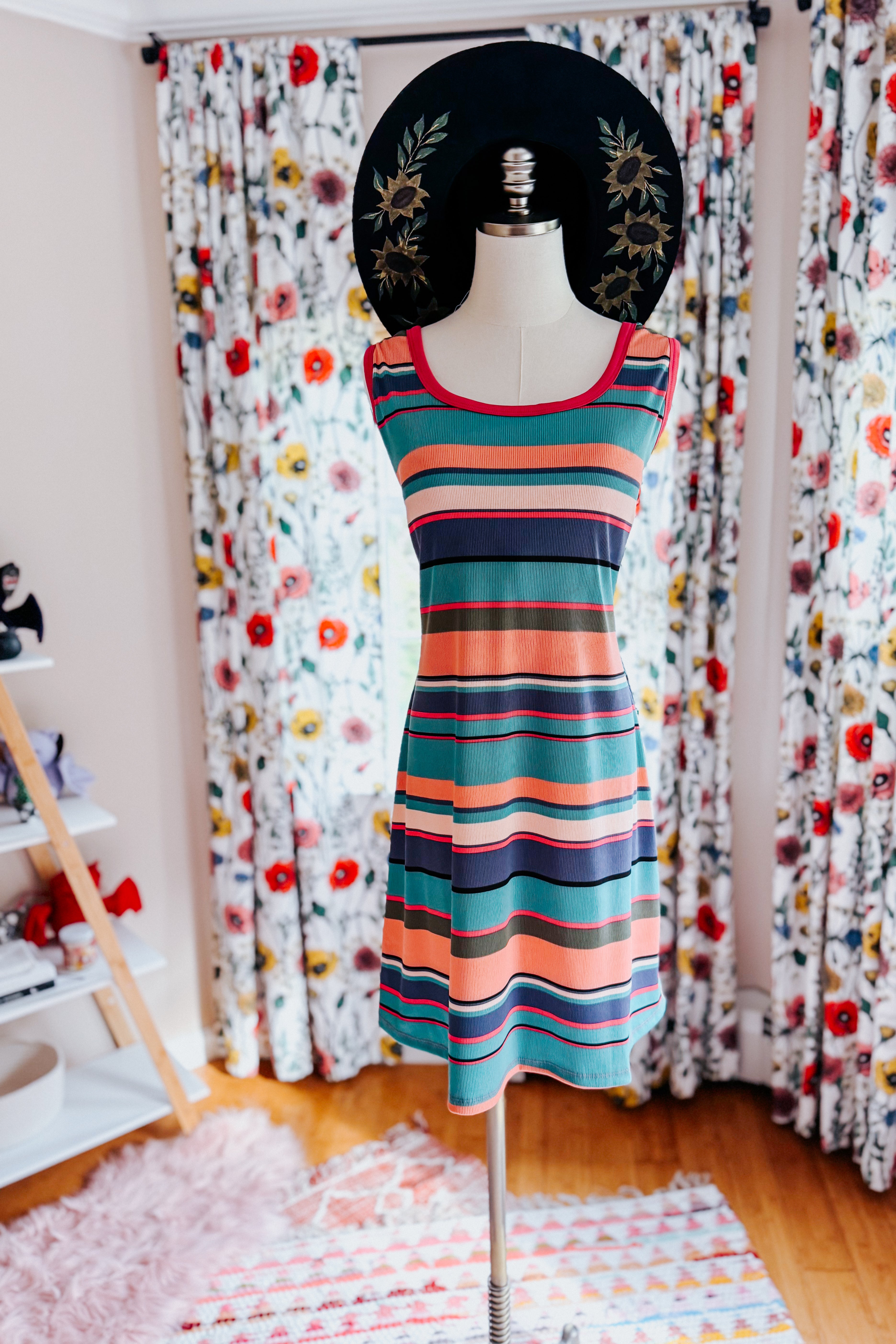 Colorful striped tank dress displayed on mannequin with wide black floral hat in cozy room with floral curtains