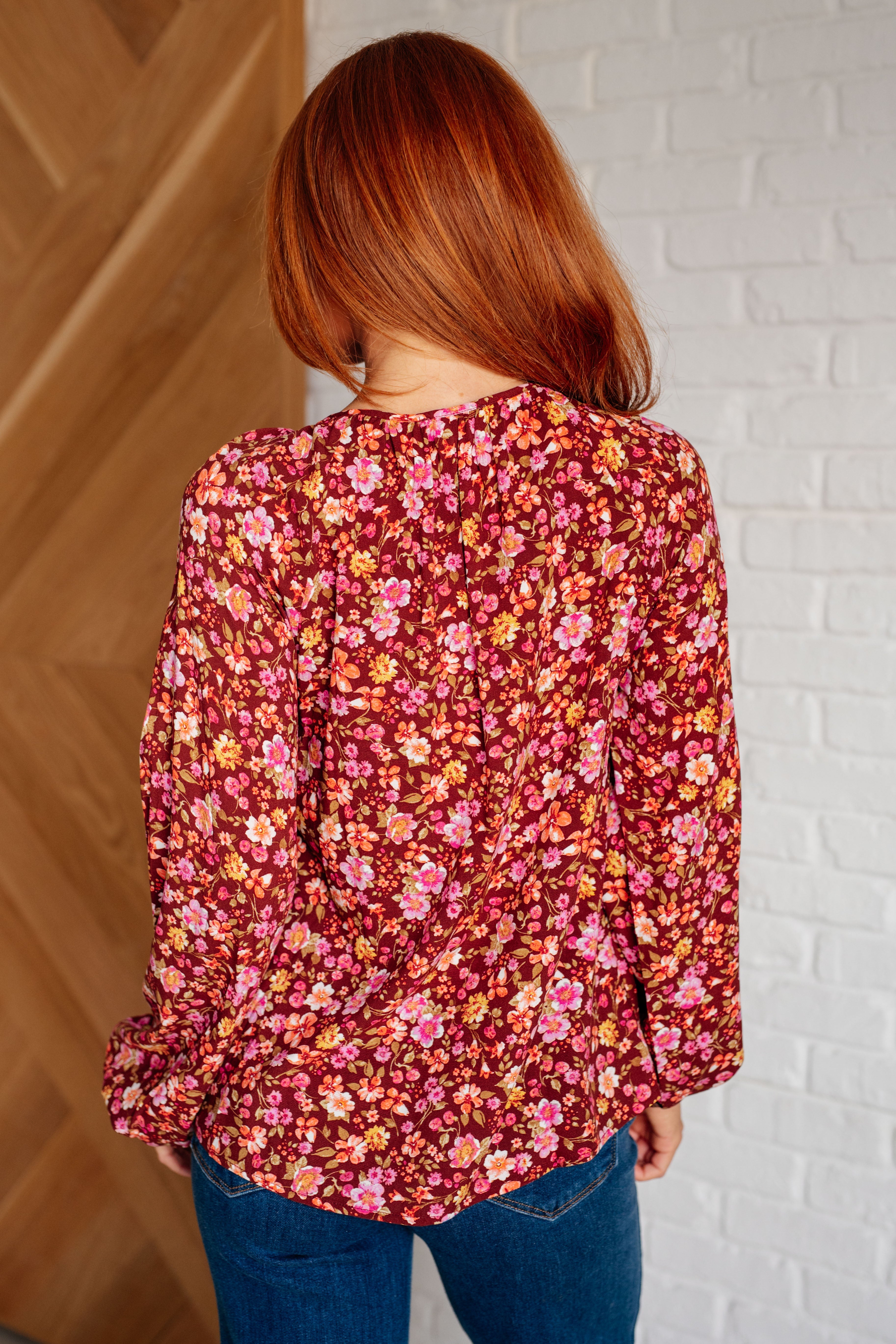 Woman wearing a rust floral blouse with pink and orange flowers paired with blue jeans against a white brick wall