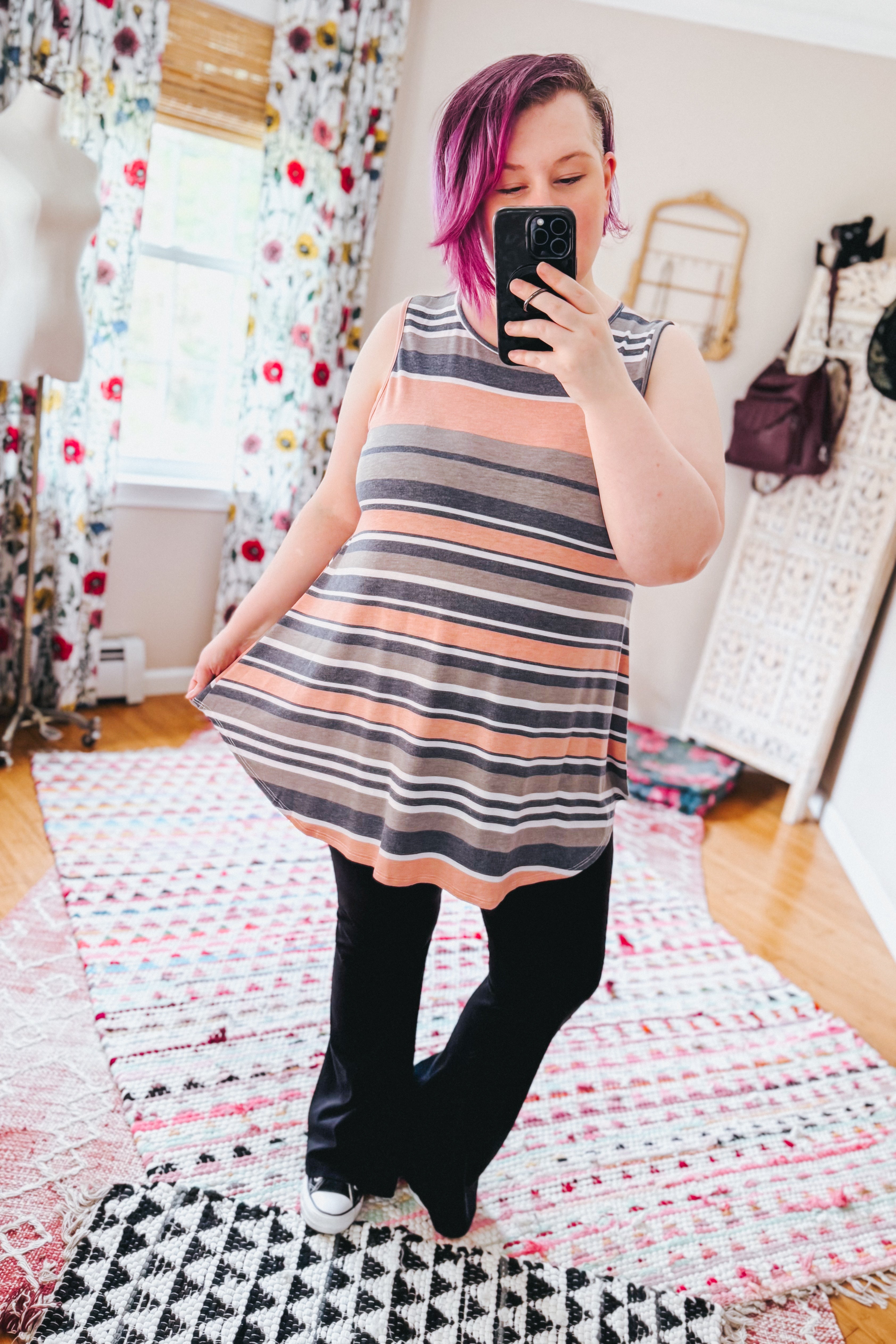 Woman wearing a sleeveless striped tunic top with shades of pink and gray paired with black pants in a cozy room