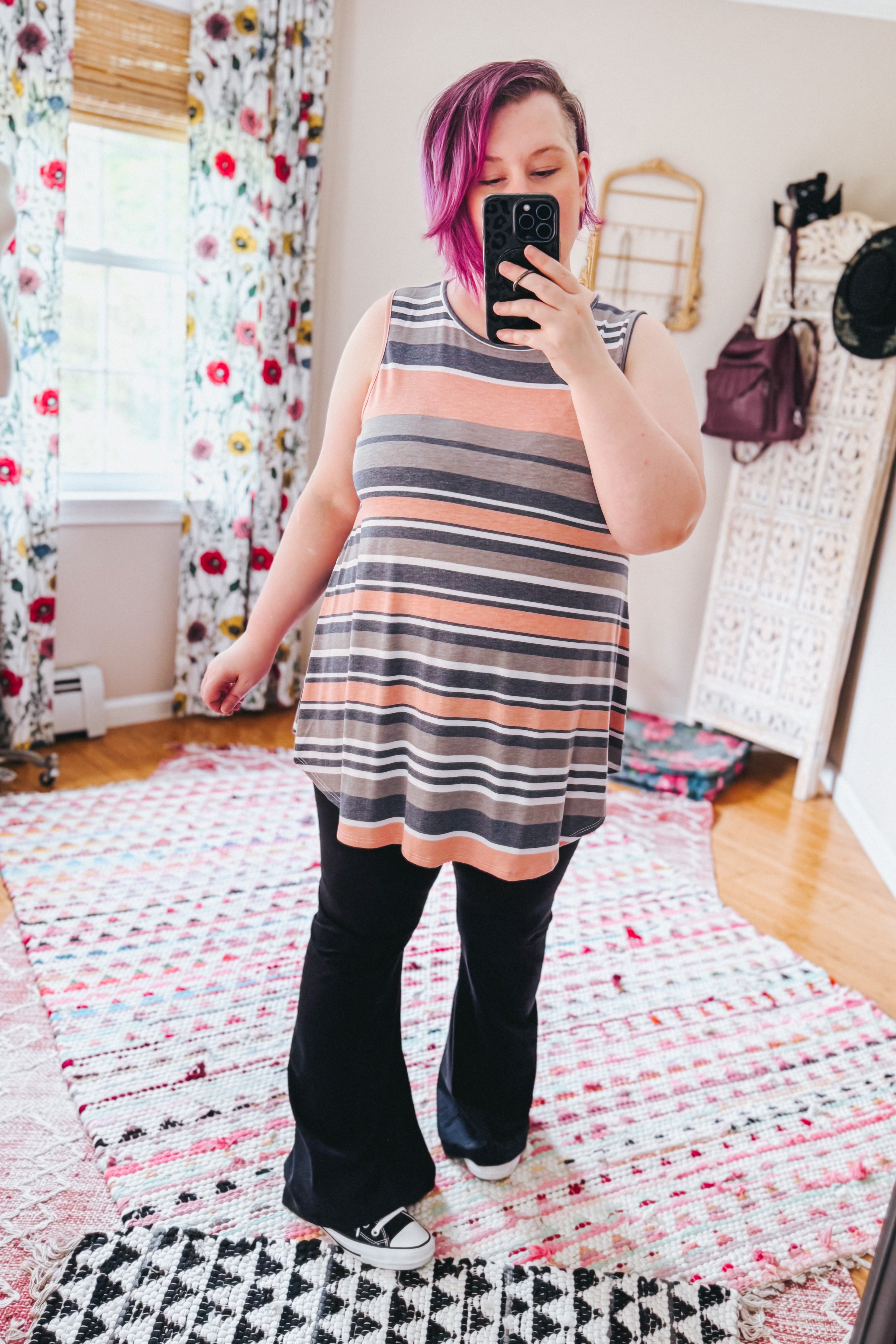 Woman with vibrant pink hair wearing a striped sleeveless top and black pants standing in a bright room with floral curtains