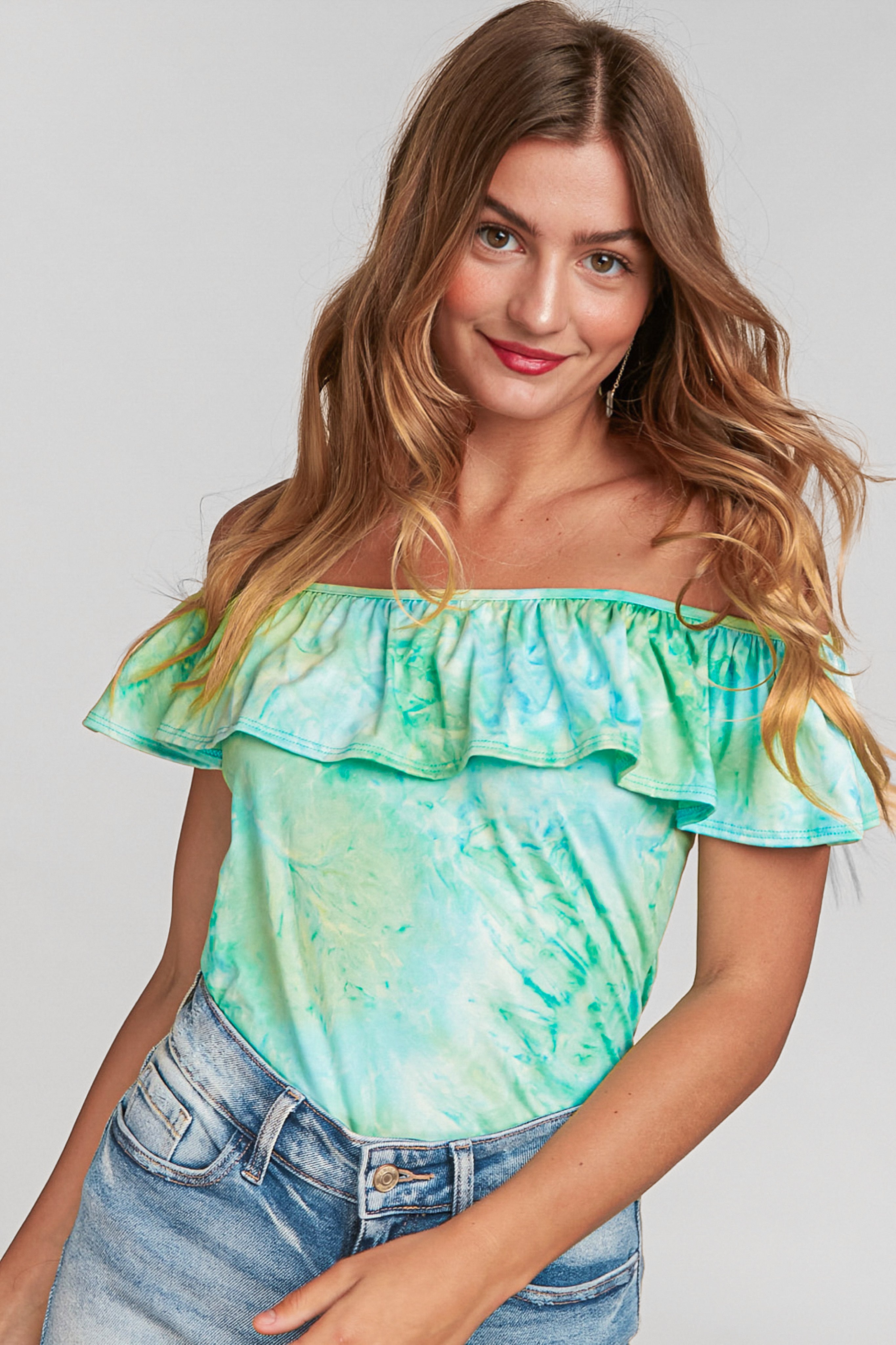 Woman with loose wavy hair wearing blue-green tie dye off-shoulder ruffle top and denim shorts smiling confidently