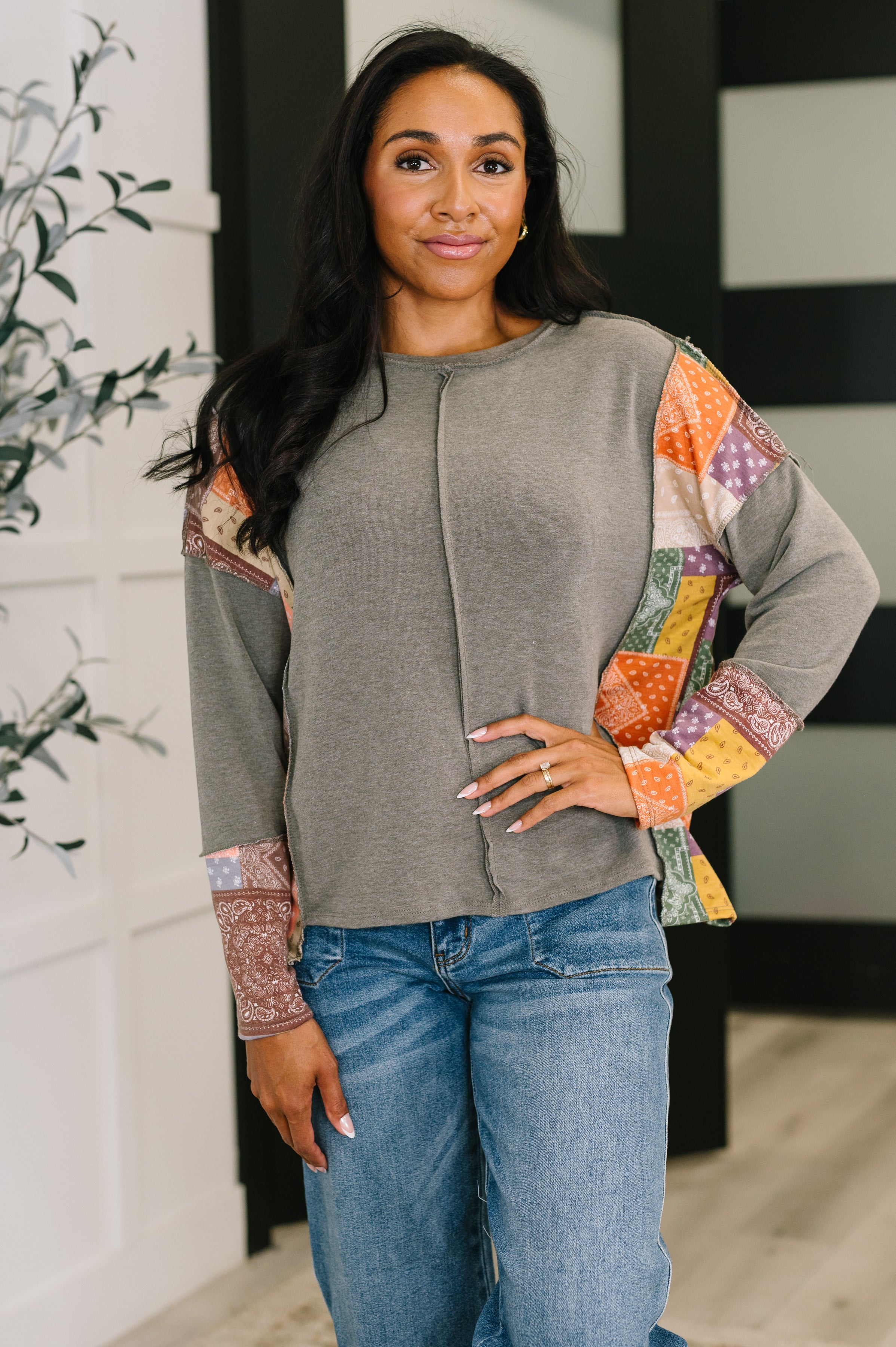 Woman wearing an olive relaxed long sleeve with colorful patchwork sleeves paired with blue denim jeans indoors