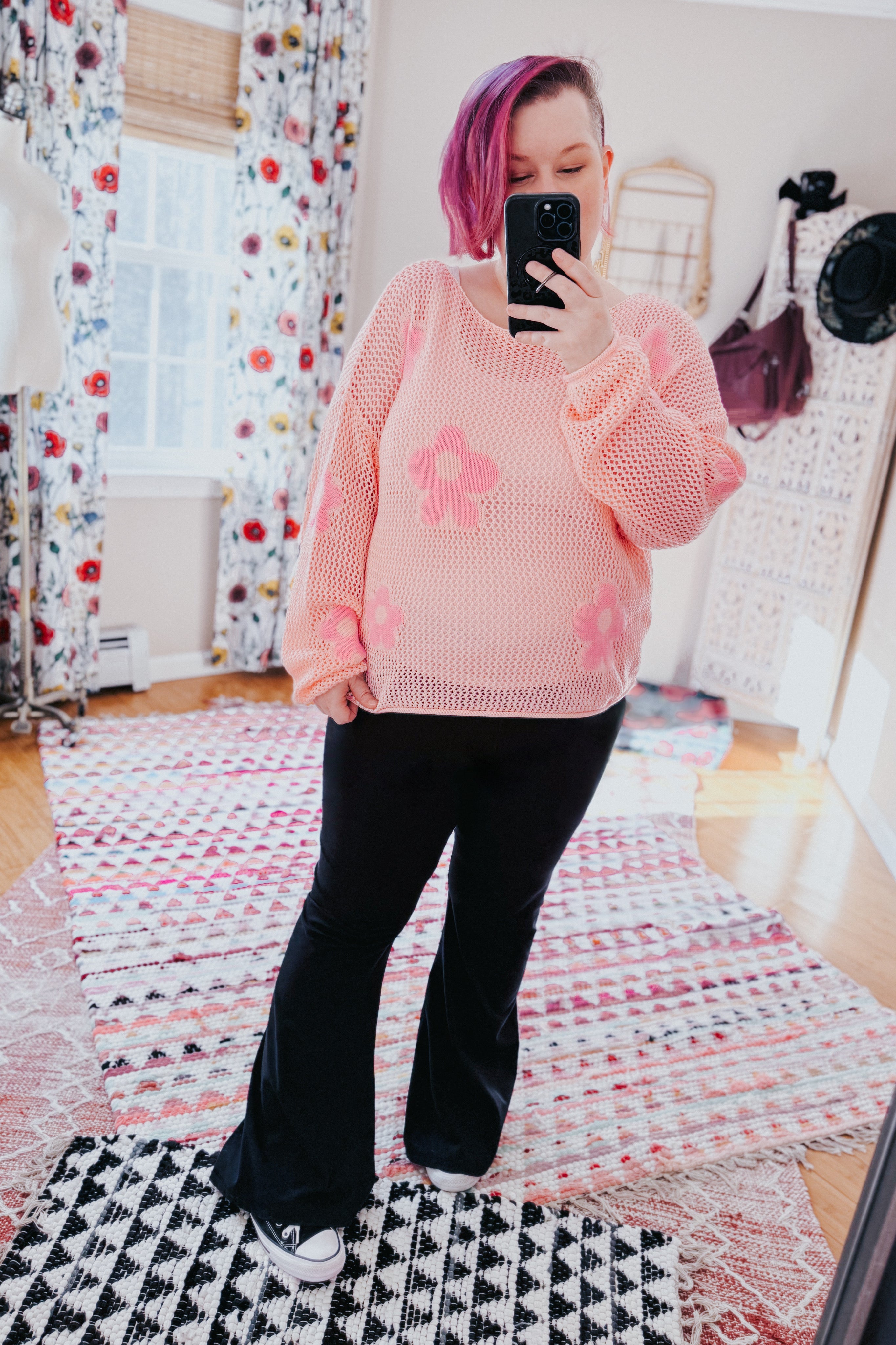 Woman wearing a blush pink floral mesh sweater top paired with black flared pants and sneakers indoors