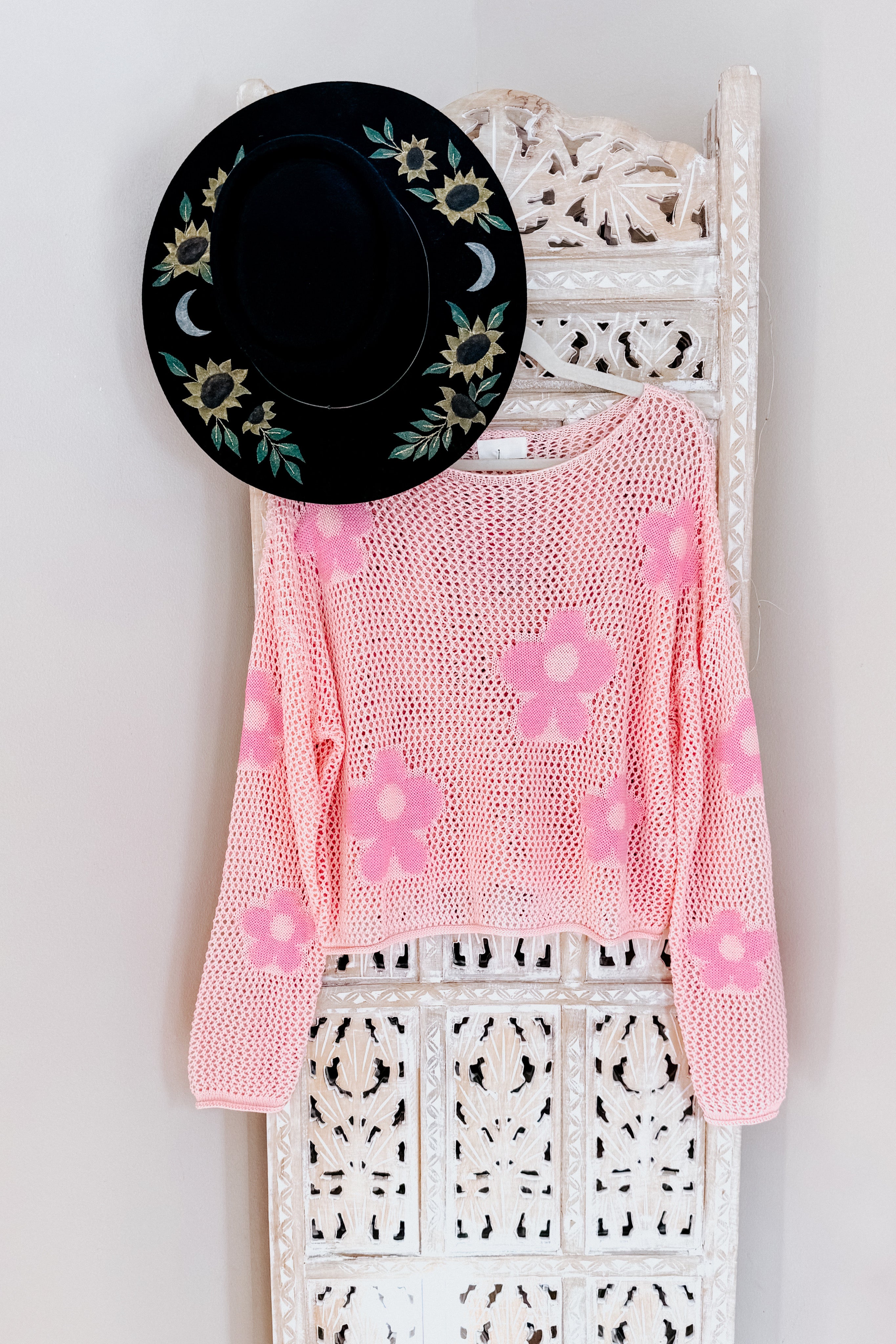 Pink floral mesh sweater top hanging on carved white wooden panel with black floral embroidered hat