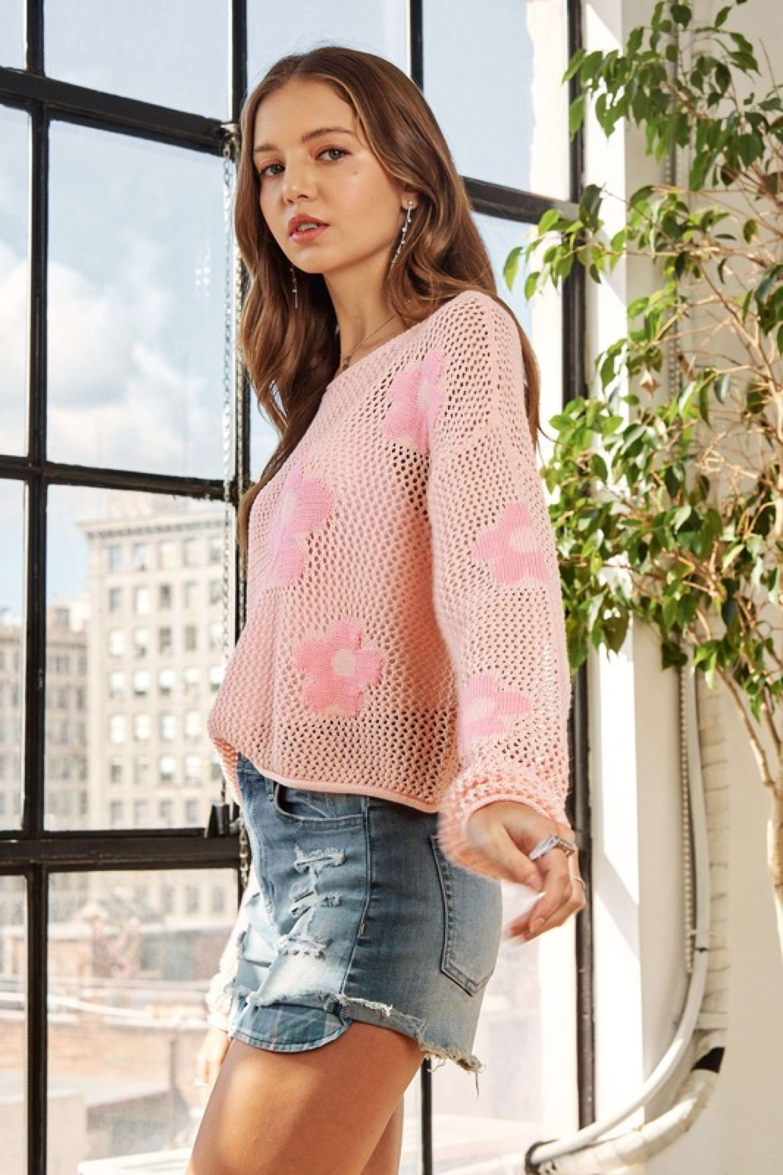 Woman wearing a pink floral mesh sweater top paired with distressed denim shorts standing indoors by a window