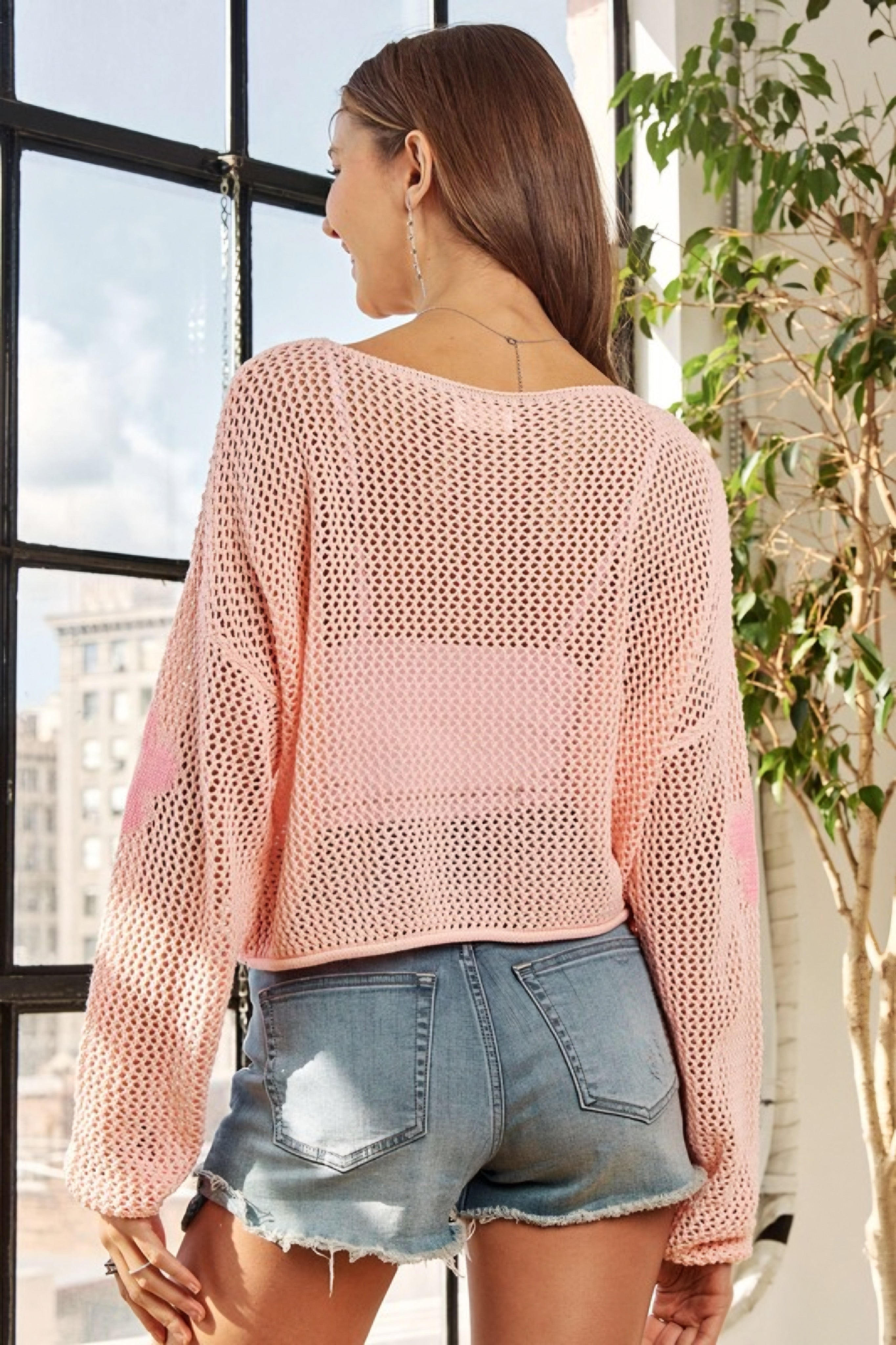 Woman seen from behind wearing a blush pink mesh sweater top and light blue denim cutoff shorts indoors by a window