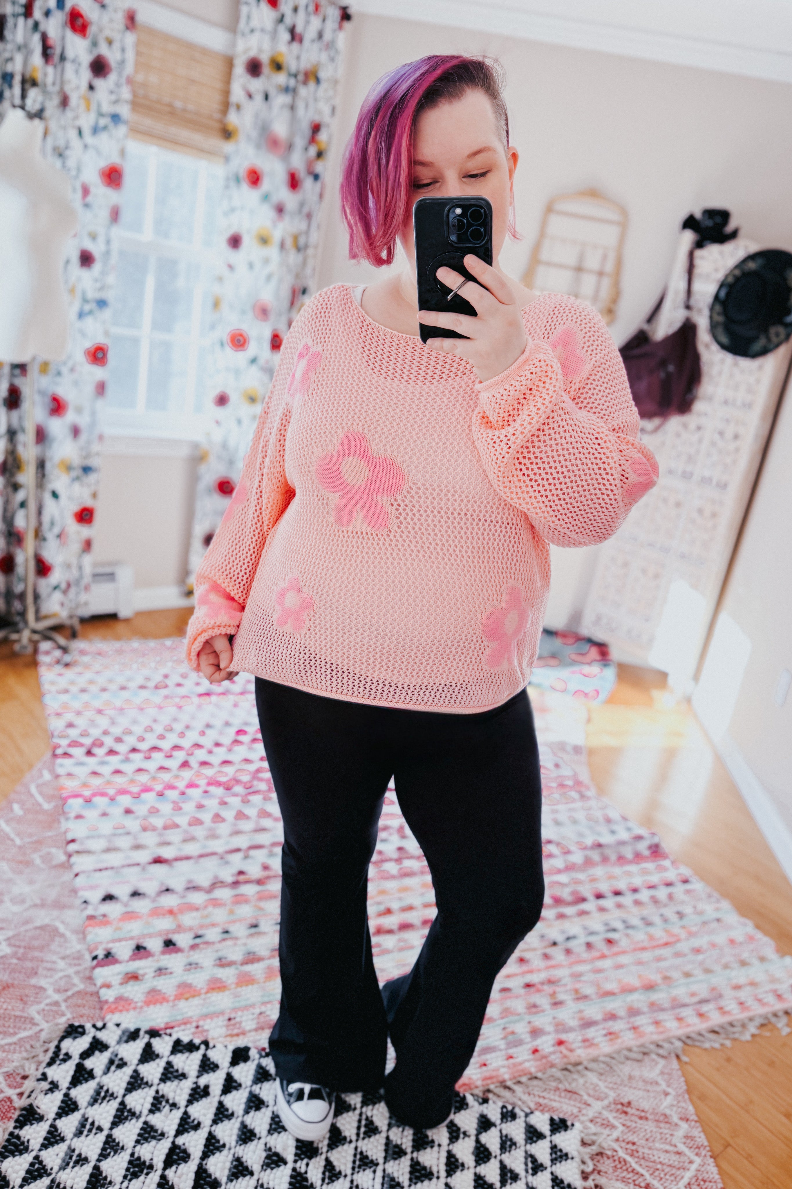 Woman wearing a blush pink floral mesh sweater with oversized sleeves and black pants, taking a mirror selfie indoors