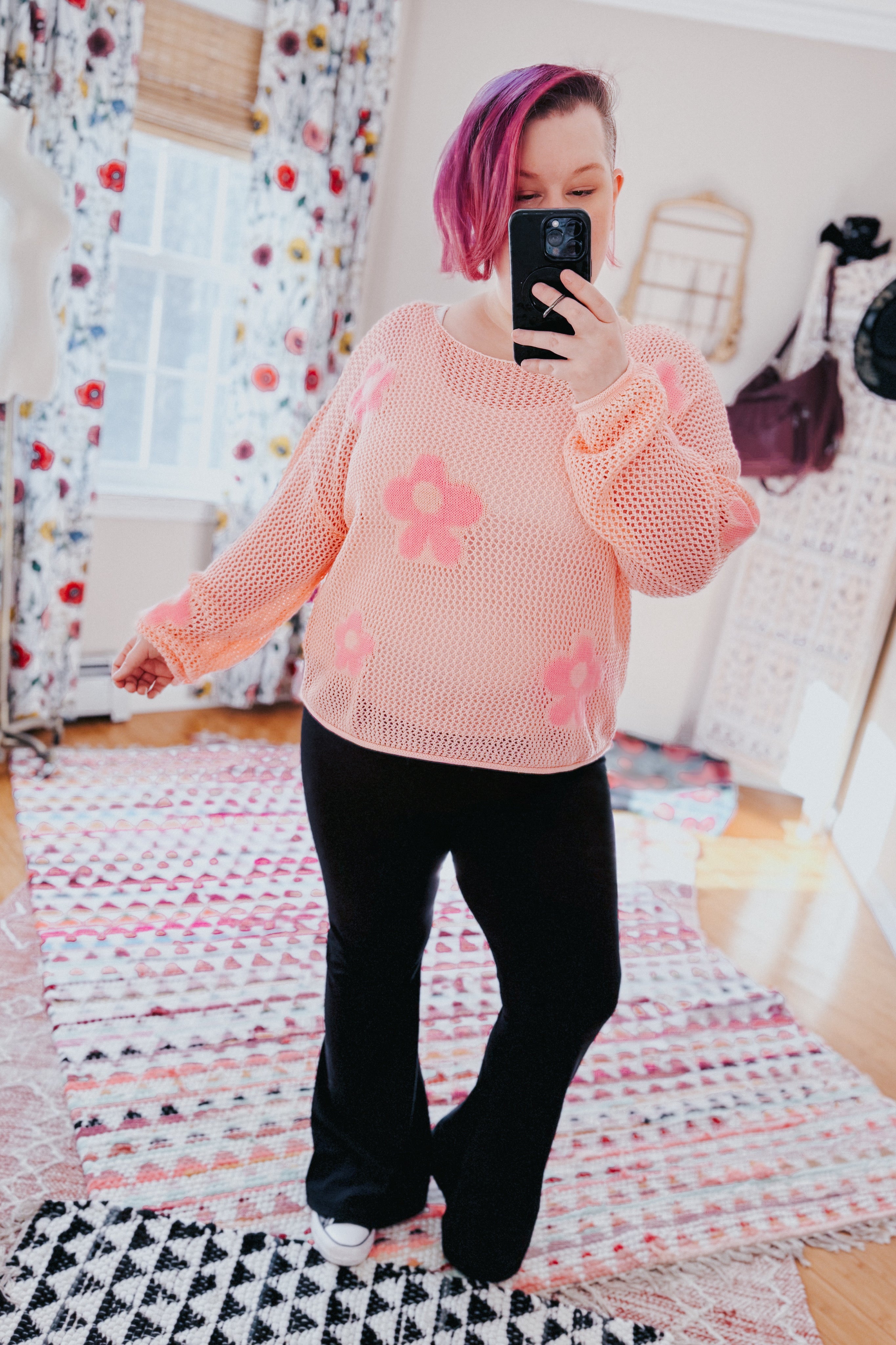 Woman wearing a blush pink mesh sweater with floral accents paired with black flared pants indoors taking a selfie