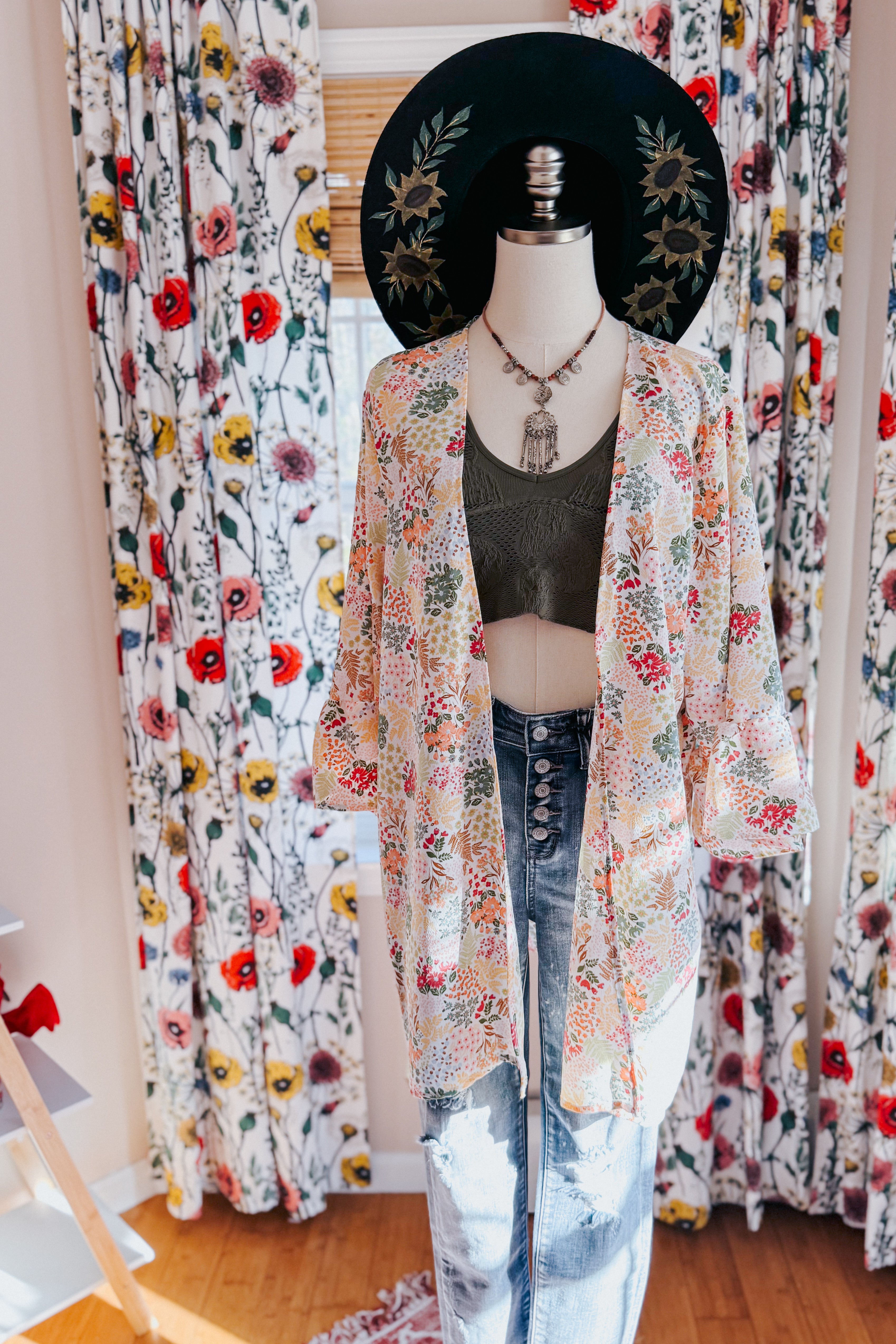 Mannequin wearing ivory floral light cardigan over olive crop top and high-waisted button fly jeans with black hat indoors