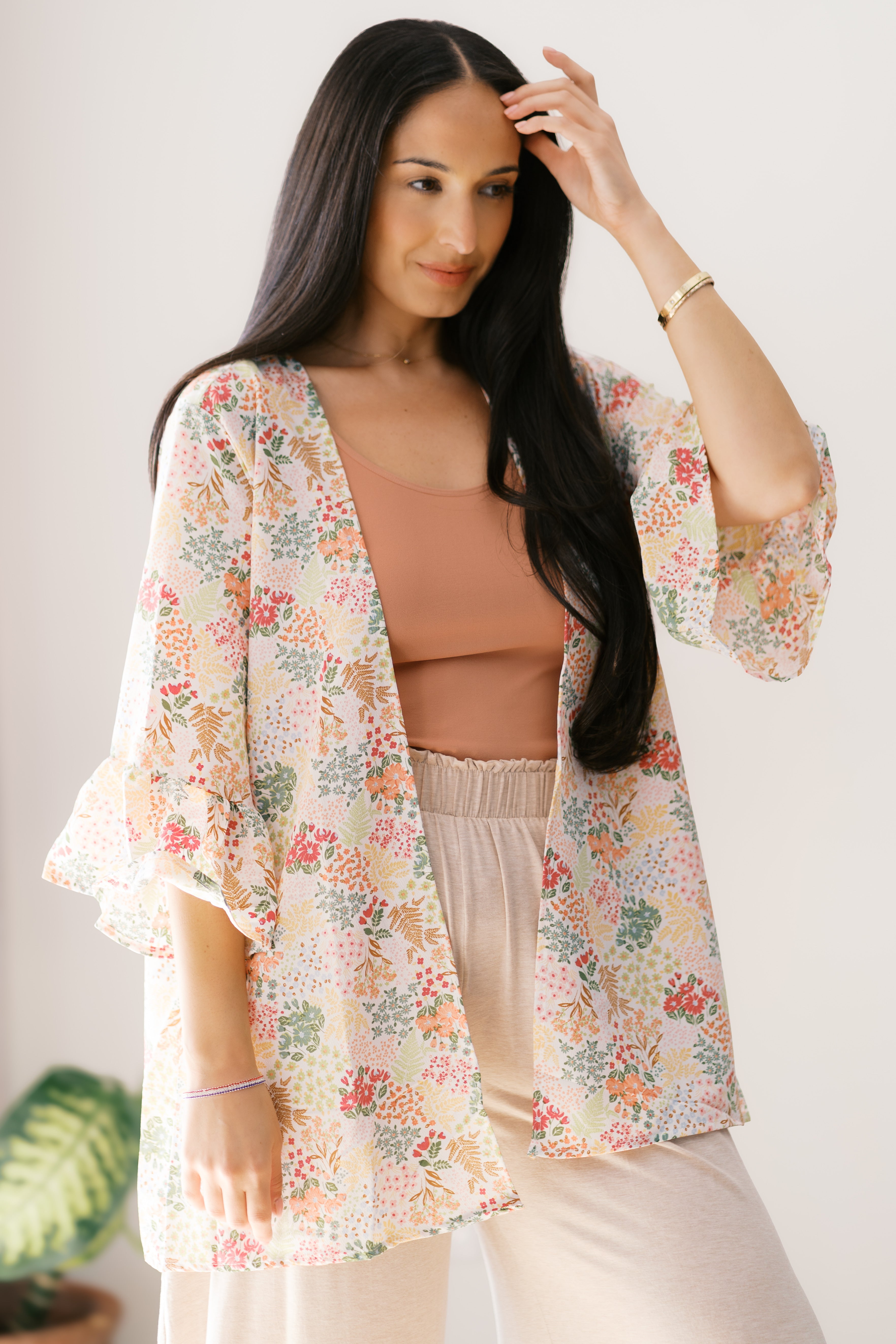 Woman wearing an ivory floral light cardigan with ruffle sleeves over a rust tank and cream pants indoors