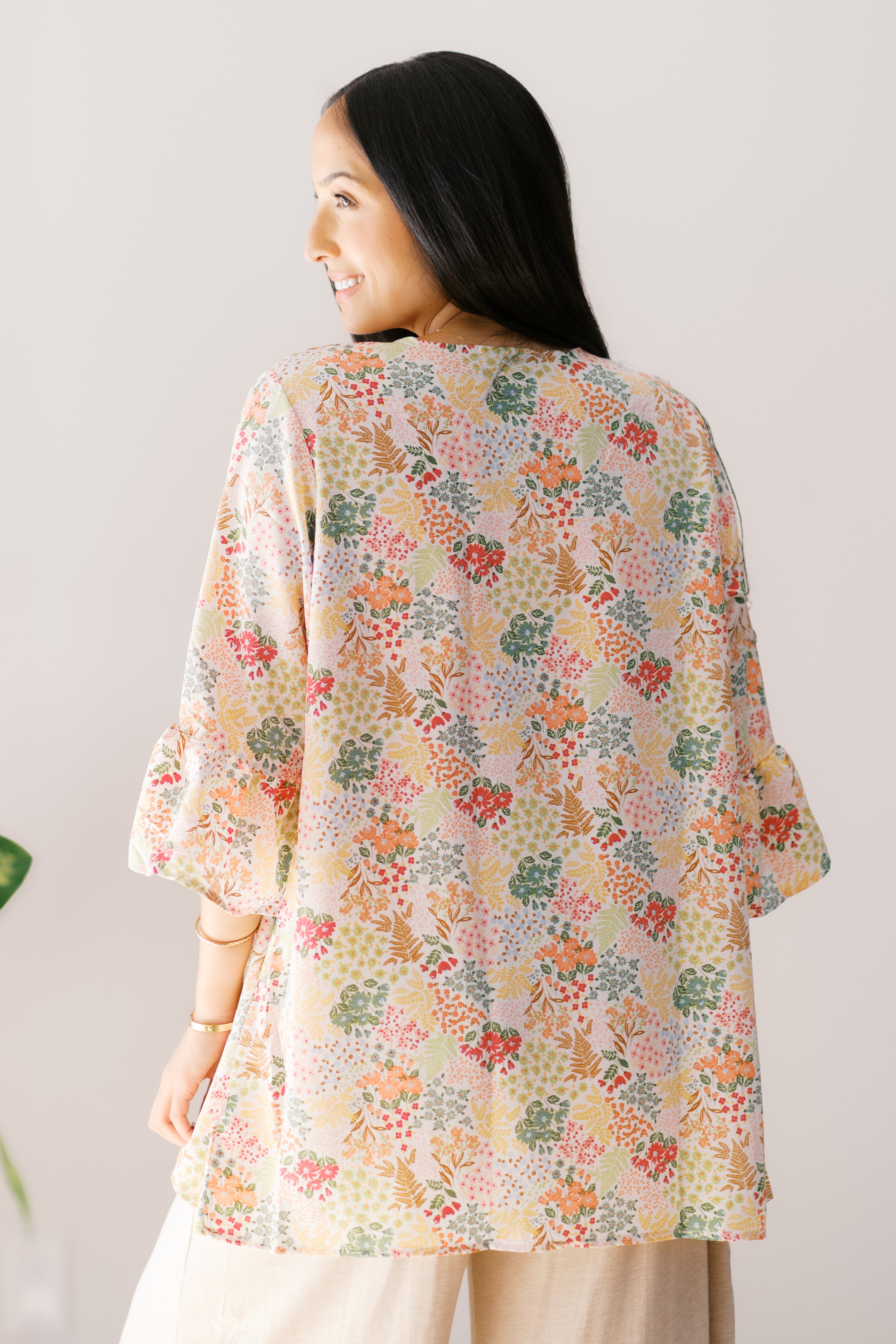 Smiling woman wearing an ivory floral Sweet Bloom cardigan with ruffled sleeves and loose fit viewed from behind