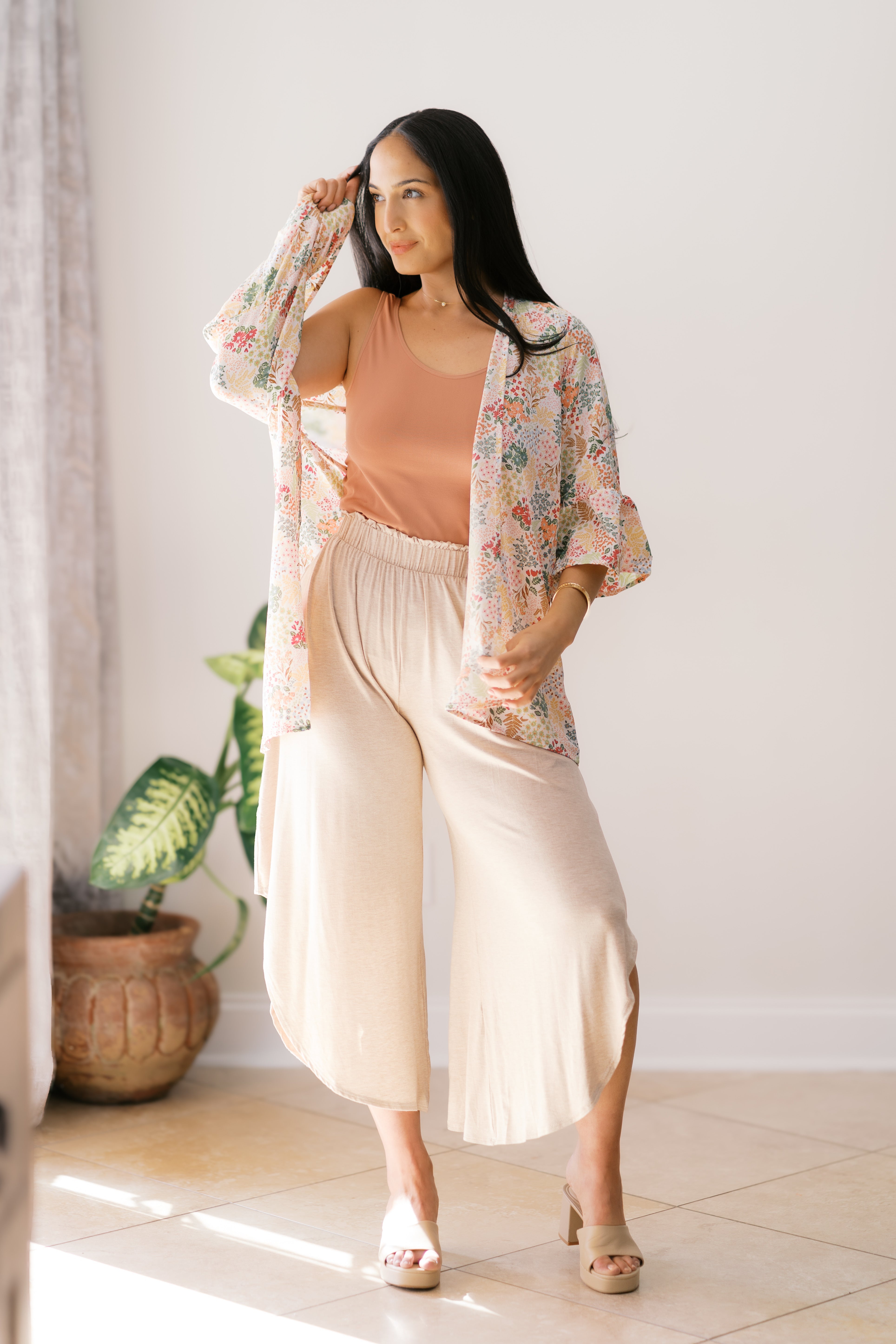 Woman wearing an ivory floral light cardigan over a rust tank top with cream wide-leg pants by a window