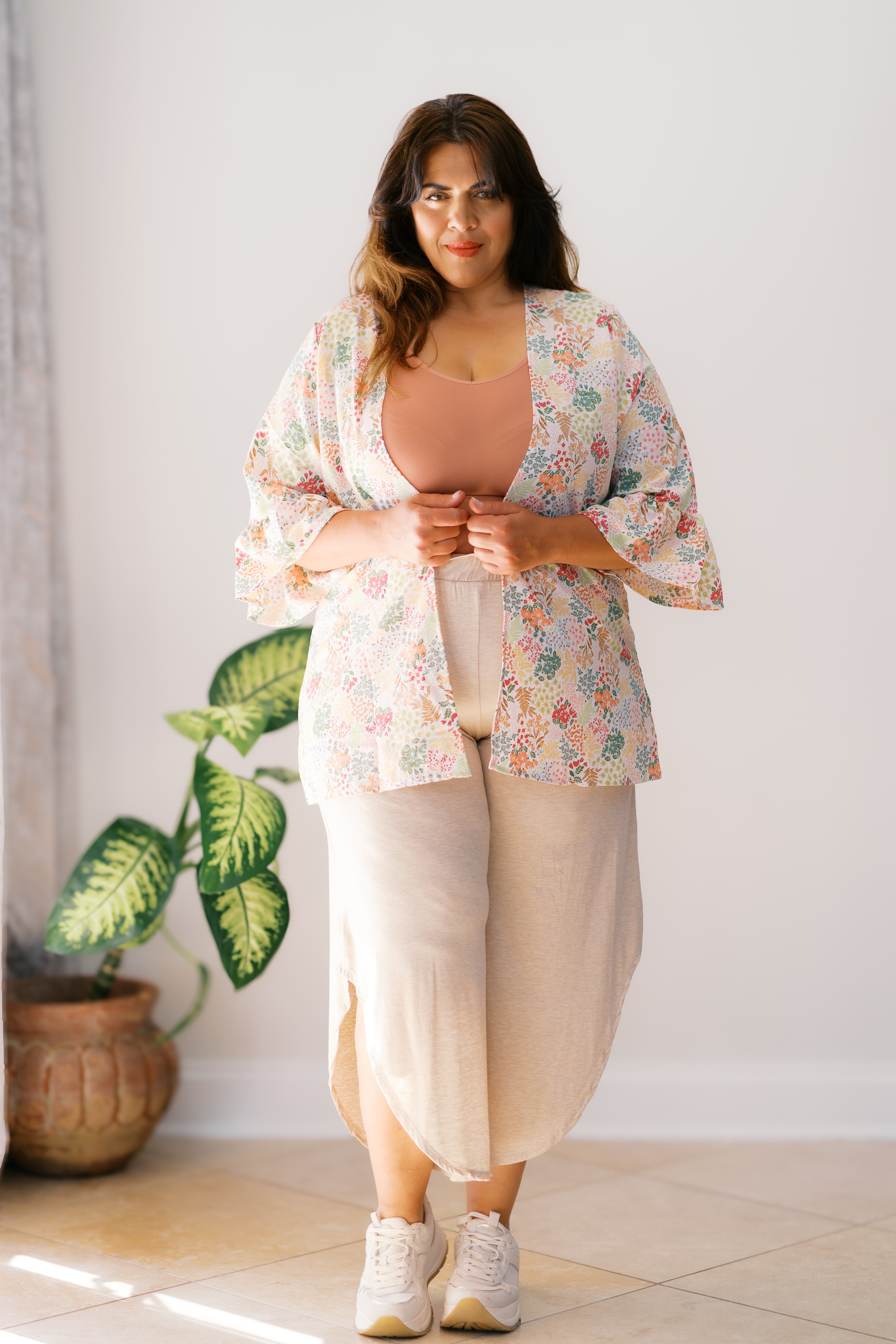 Woman wearing an ivory floral light cardigan over a rust tank top and beige skirt standing indoors by a plant