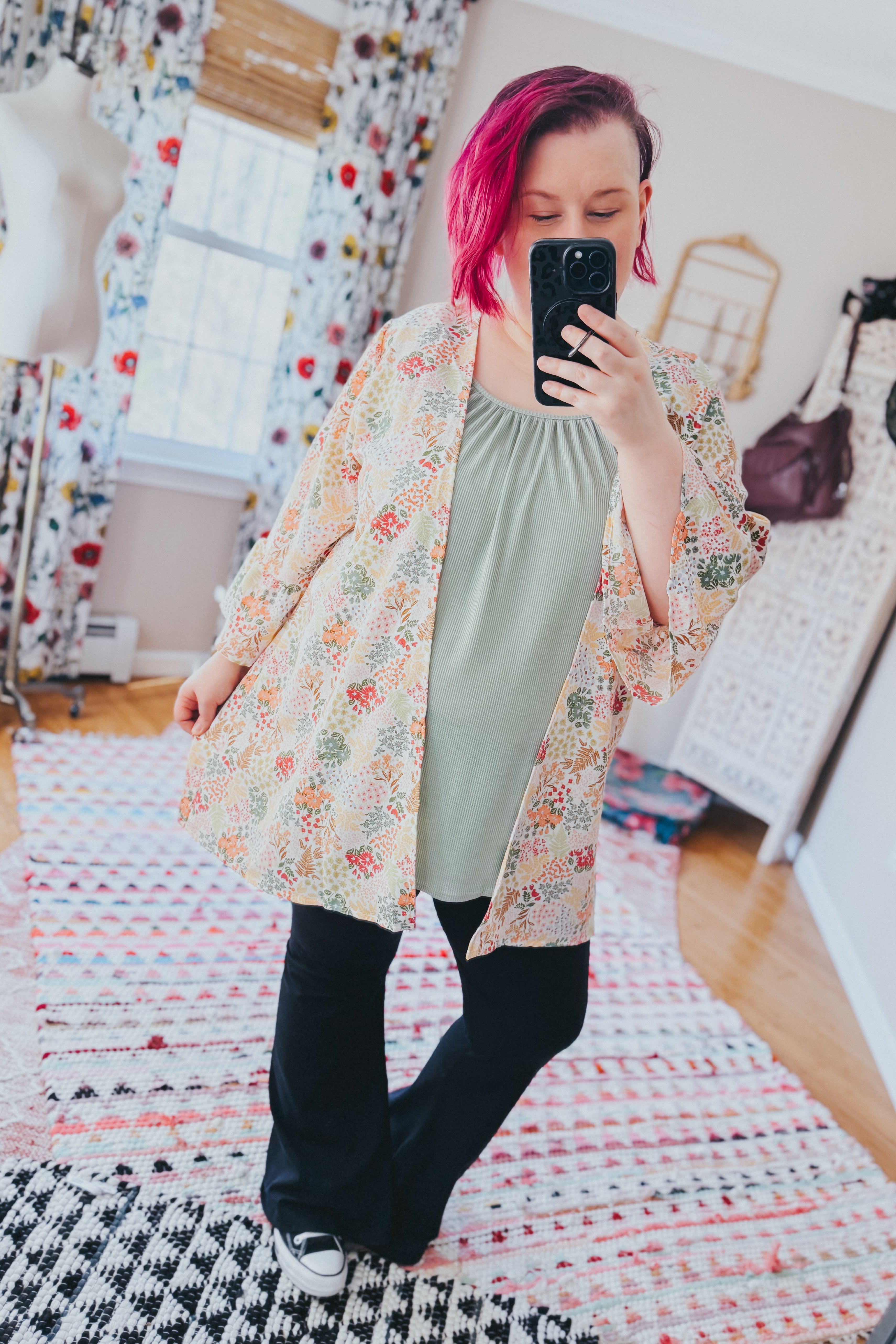 Woman with magenta hair wearing an ivory patterned light cardigan over sage top and black pants indoors taking a selfie