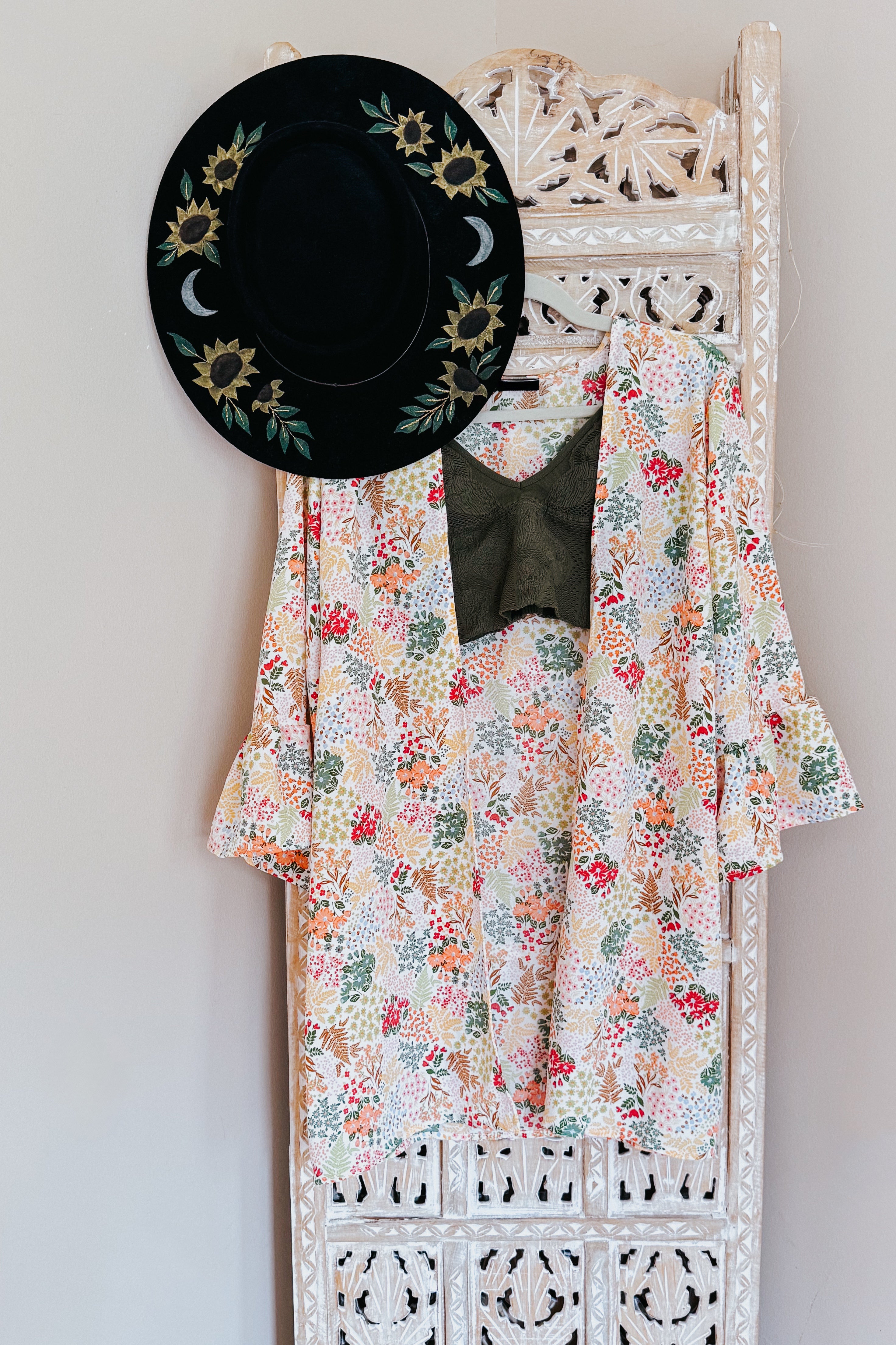Ivory floral light cardigan with ruffled sleeves paired with dark olive top and black sunflower hat on carved wooden panel