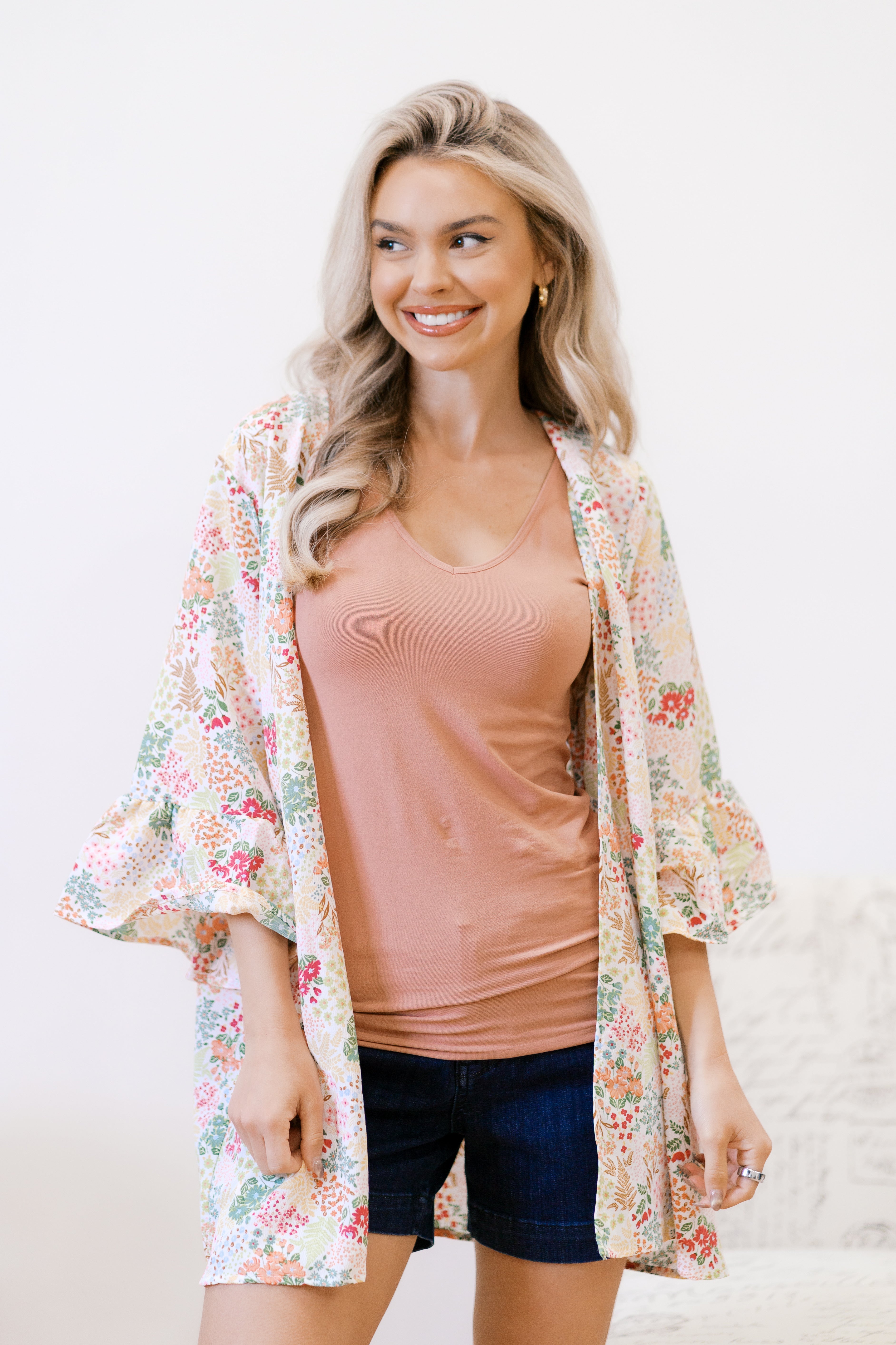 Smiling woman wearing an ivory floral light cardigan over a peach tank top and dark denim shorts indoors