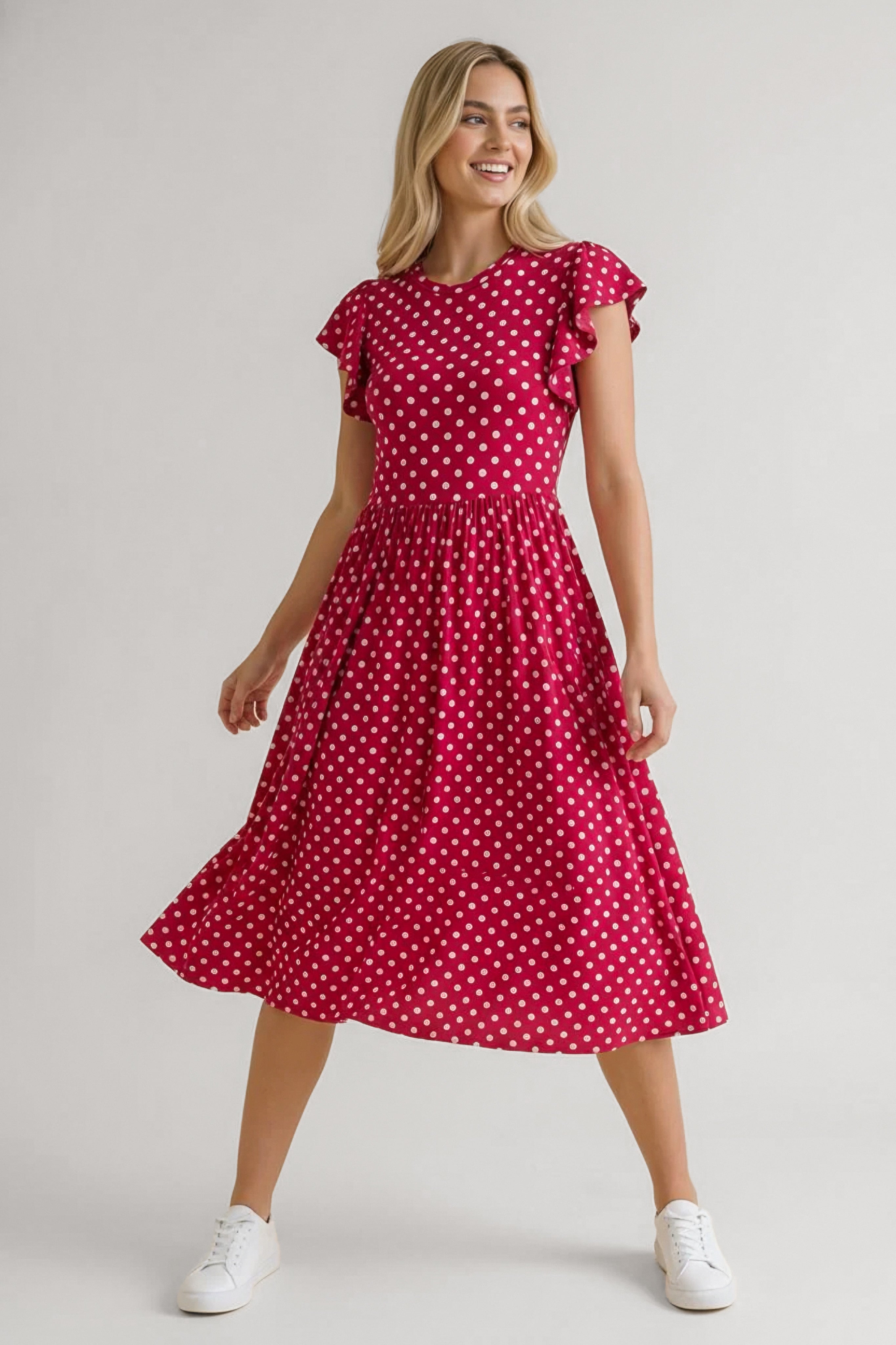 Smiling woman wearing red polka dot midi dress with flutter sleeves paired with white sneakers in playful pose