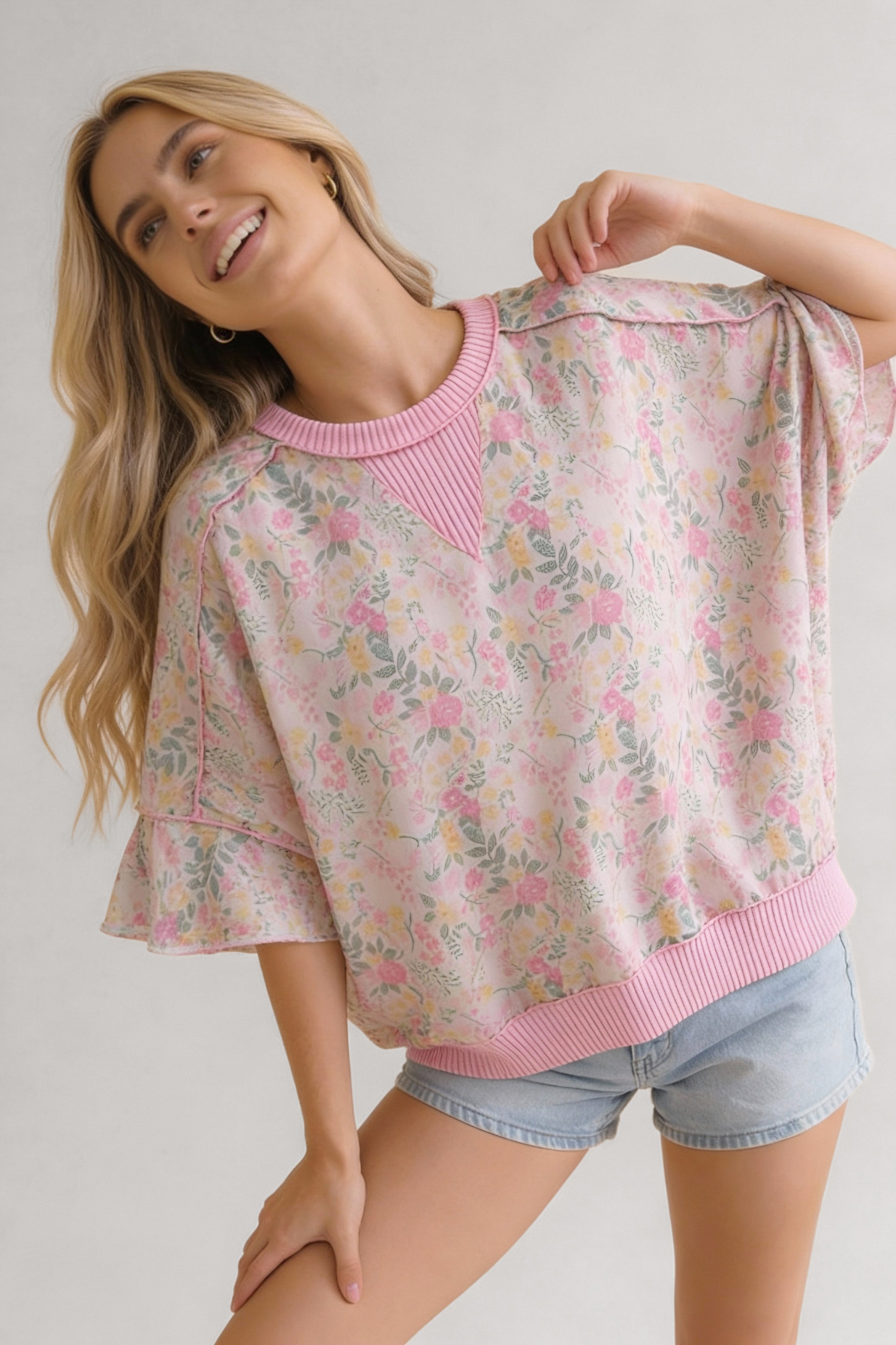 Woman smiling wearing a pink floral patterned top with ruffle sleeves and ribbed neckline and hem paired with light denim shorts