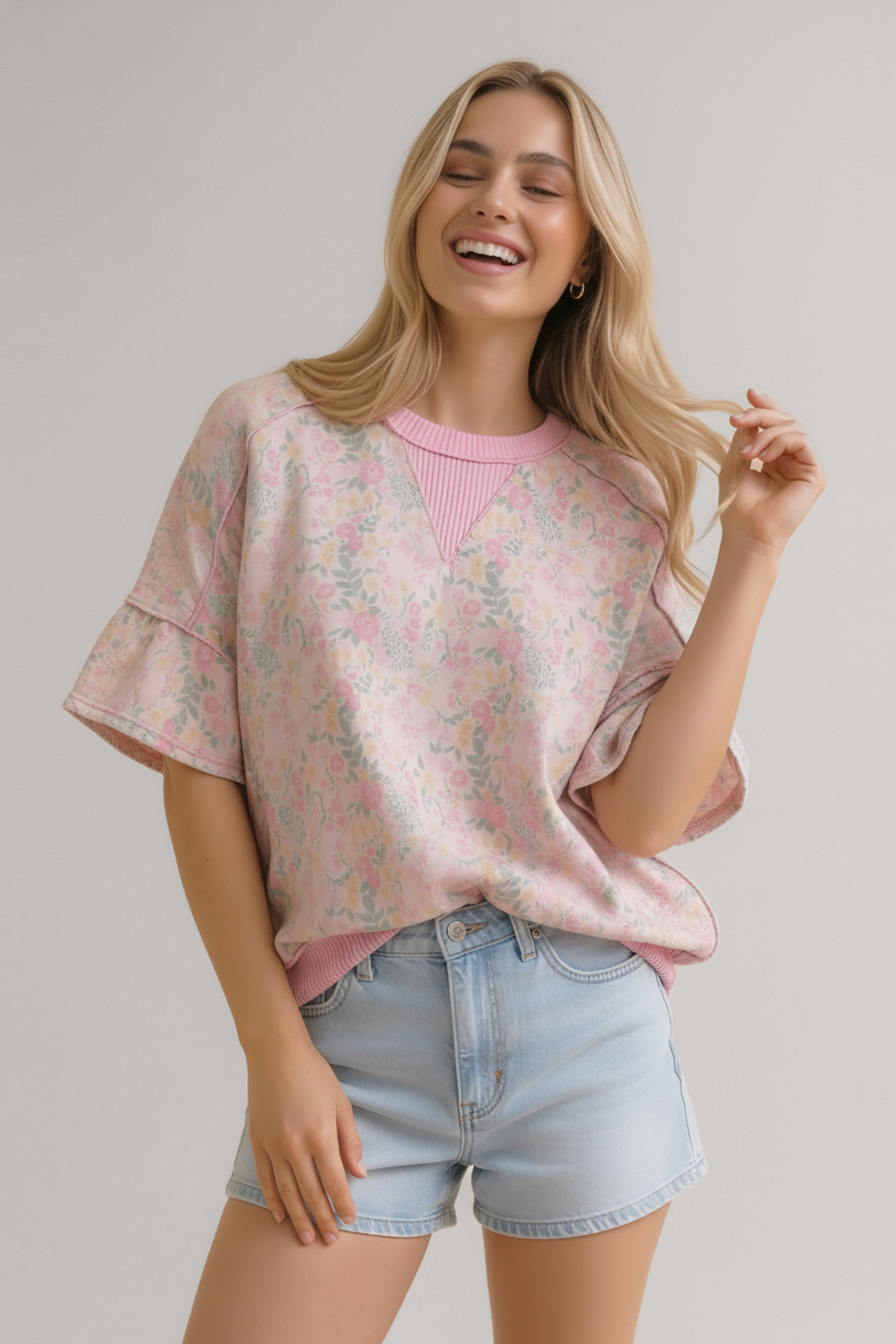 Smiling woman wearing a loose pink floral top with ruffle sleeves and ribbed neckline paired with light denim shorts