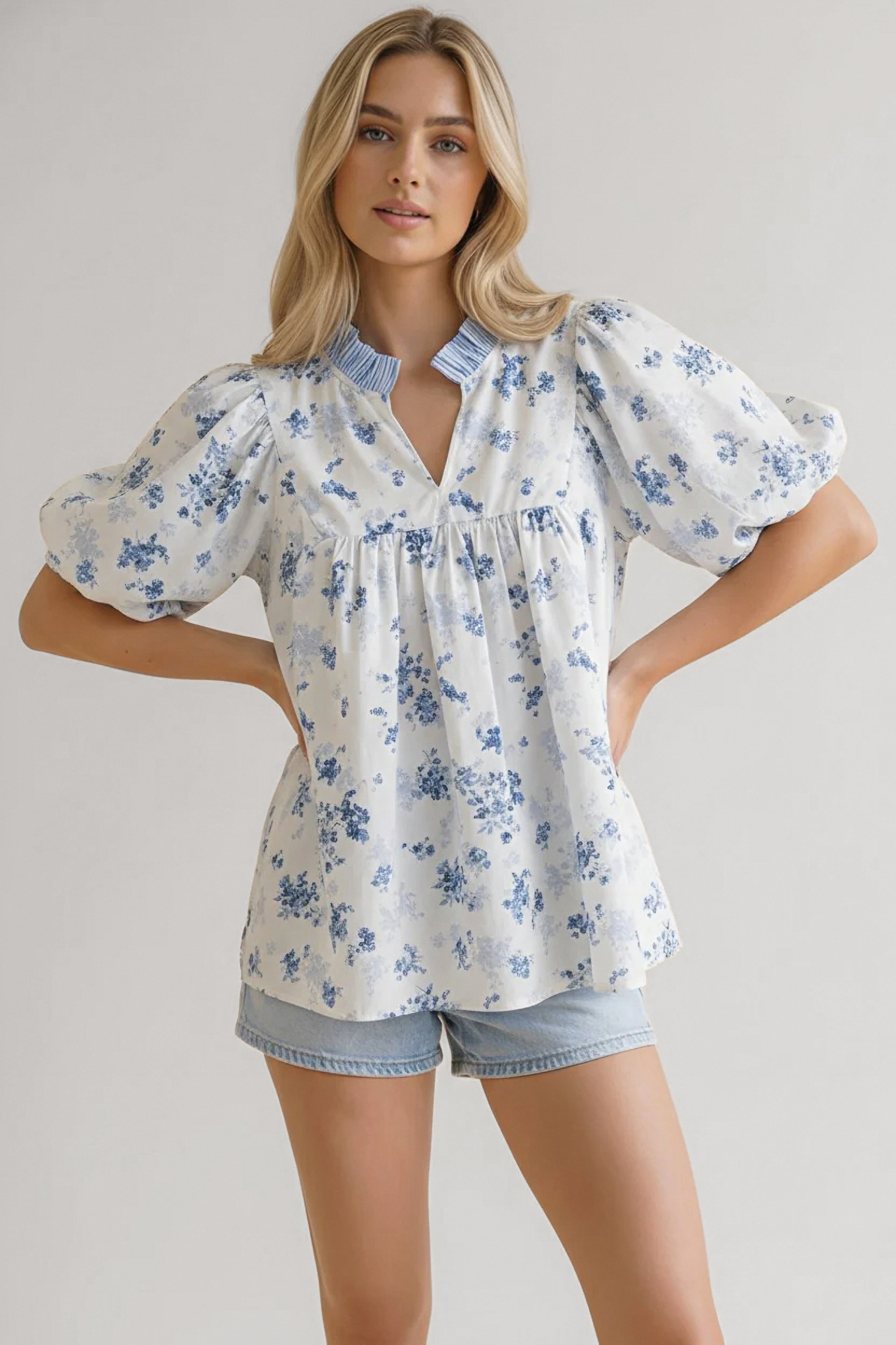 Woman wearing white blouse with blue floral print and ruffled blue collar paired with light denim shorts standing with hands on hips