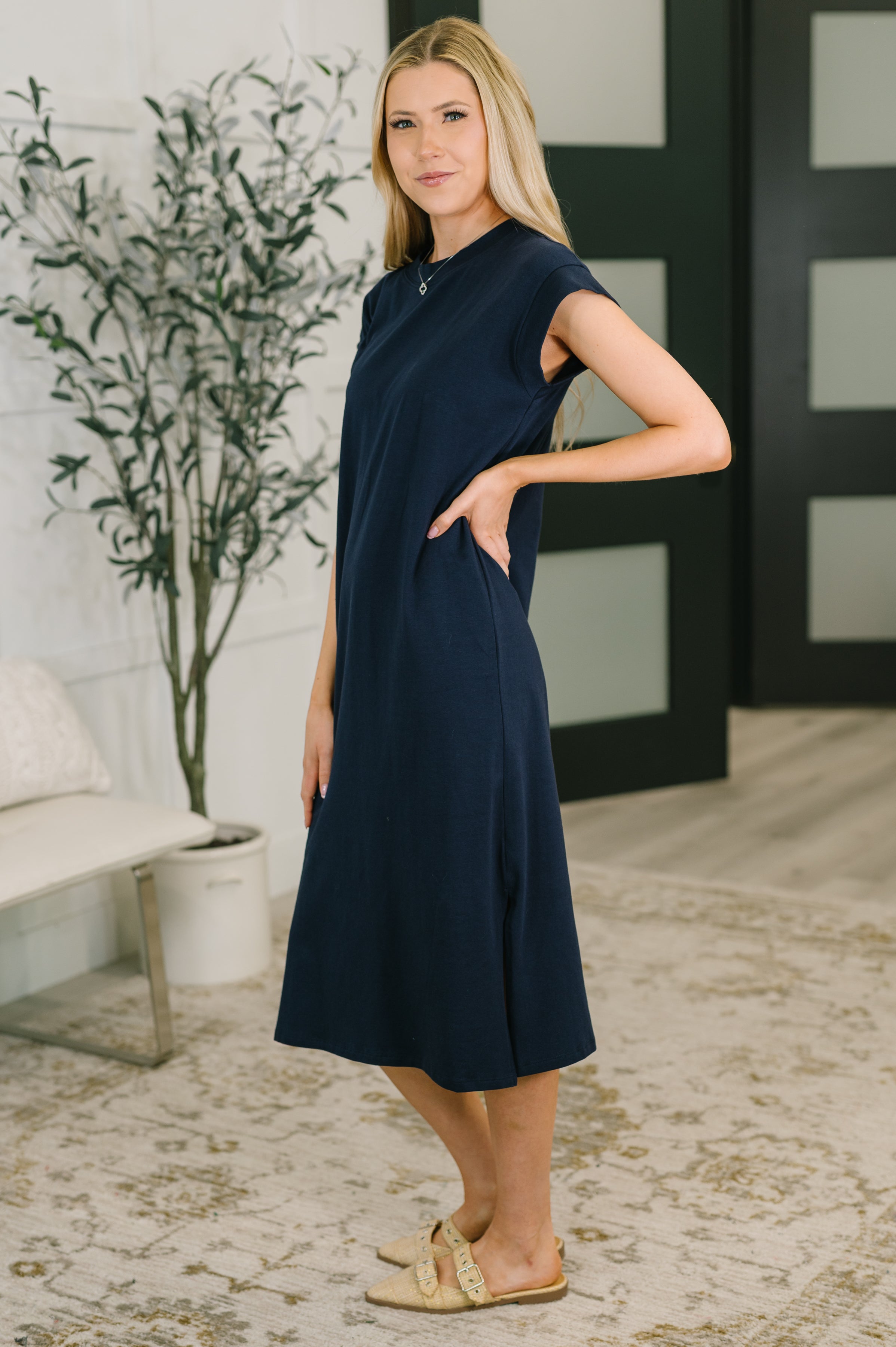 Woman wearing navy sleeveless casual midi dress standing indoors with hand on hip by plant and modern doors