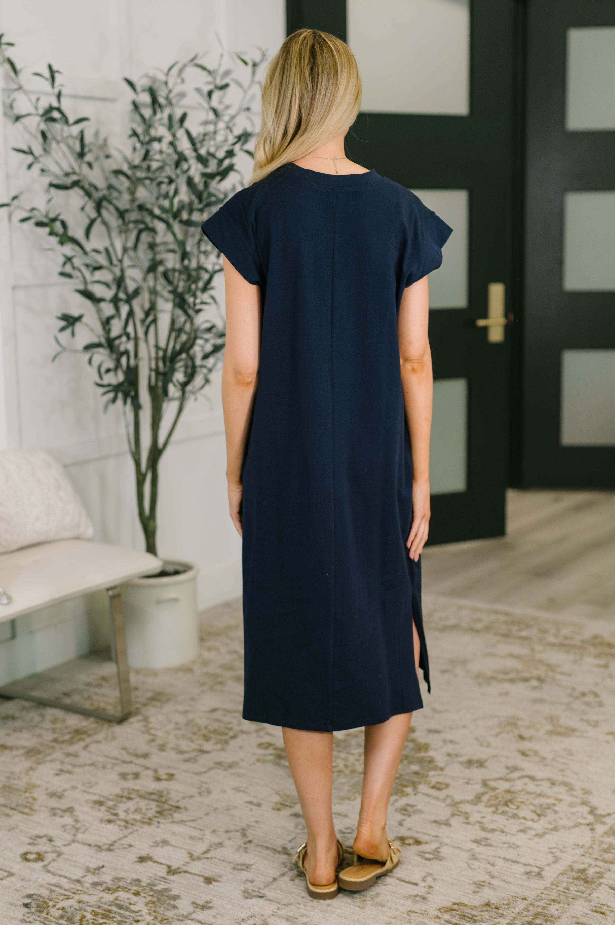 Back view of woman wearing navy casual dress with side slit and tan slide sandals standing indoors by plant and bench