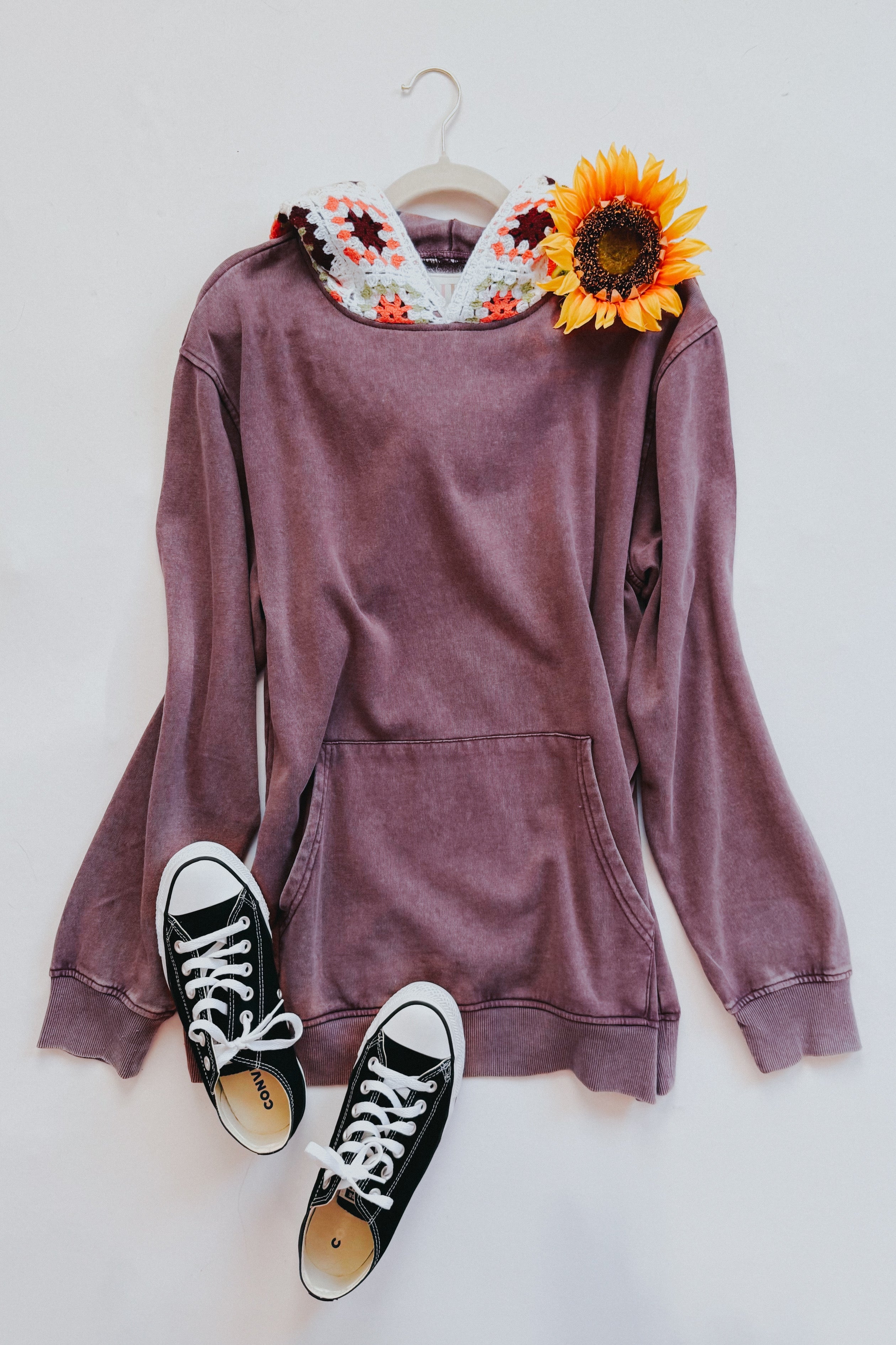 Purple acid wash hoodie with crocheted hood and sunflower on hanger paired with black canvas sneakers on white background