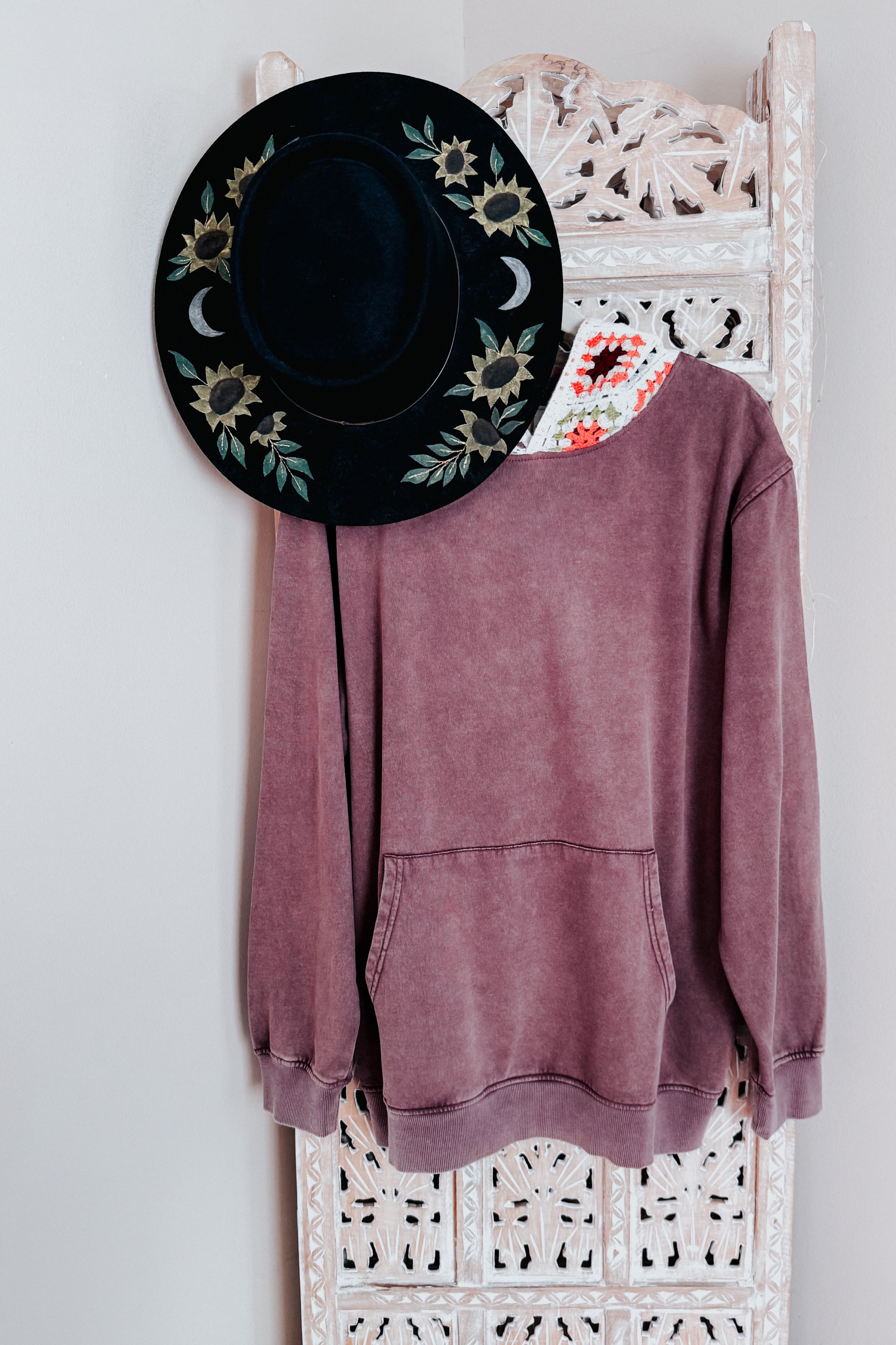 Purple acid wash hoodie with kangaroo pocket hanging on an ornate white wooden panel with a black floral hat above
