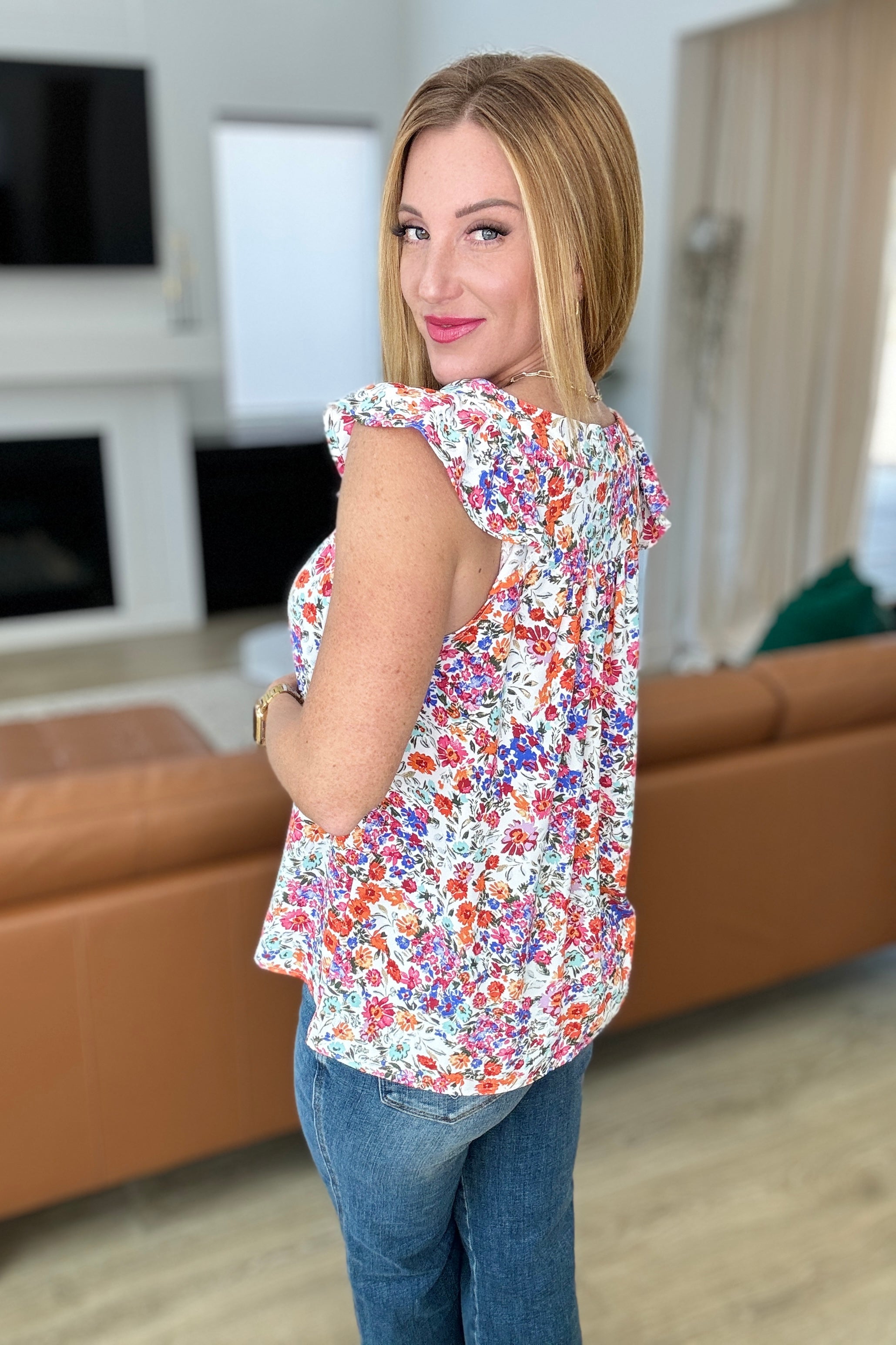 Woman wearing a colorful floral sleeveless top with ruffled shoulders and blue jeans in a cozy living room setting