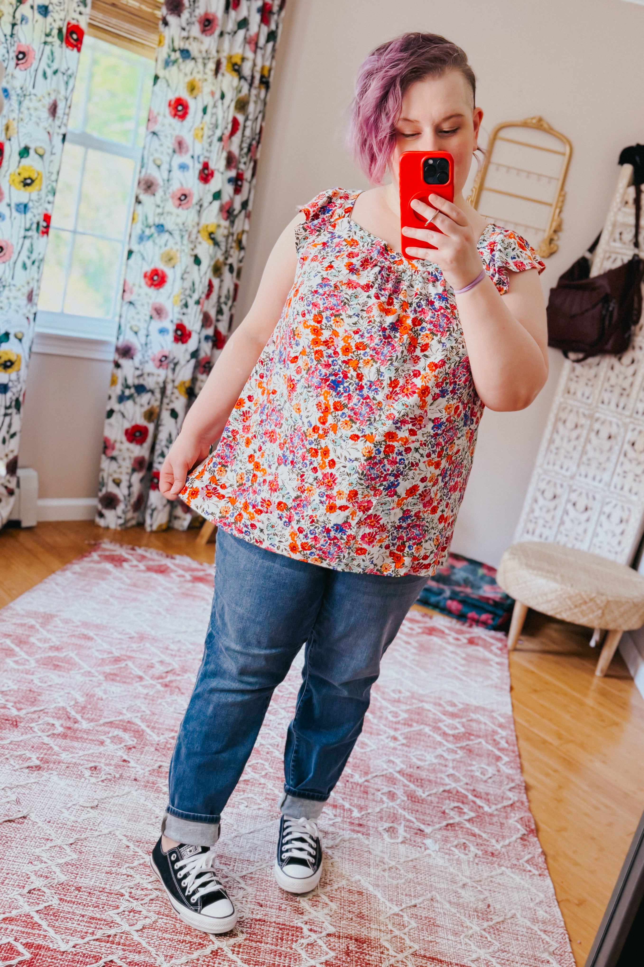 Woman wearing colorful floral red square neck top paired with cuffed jeans and black sneakers indoors in eclectic room