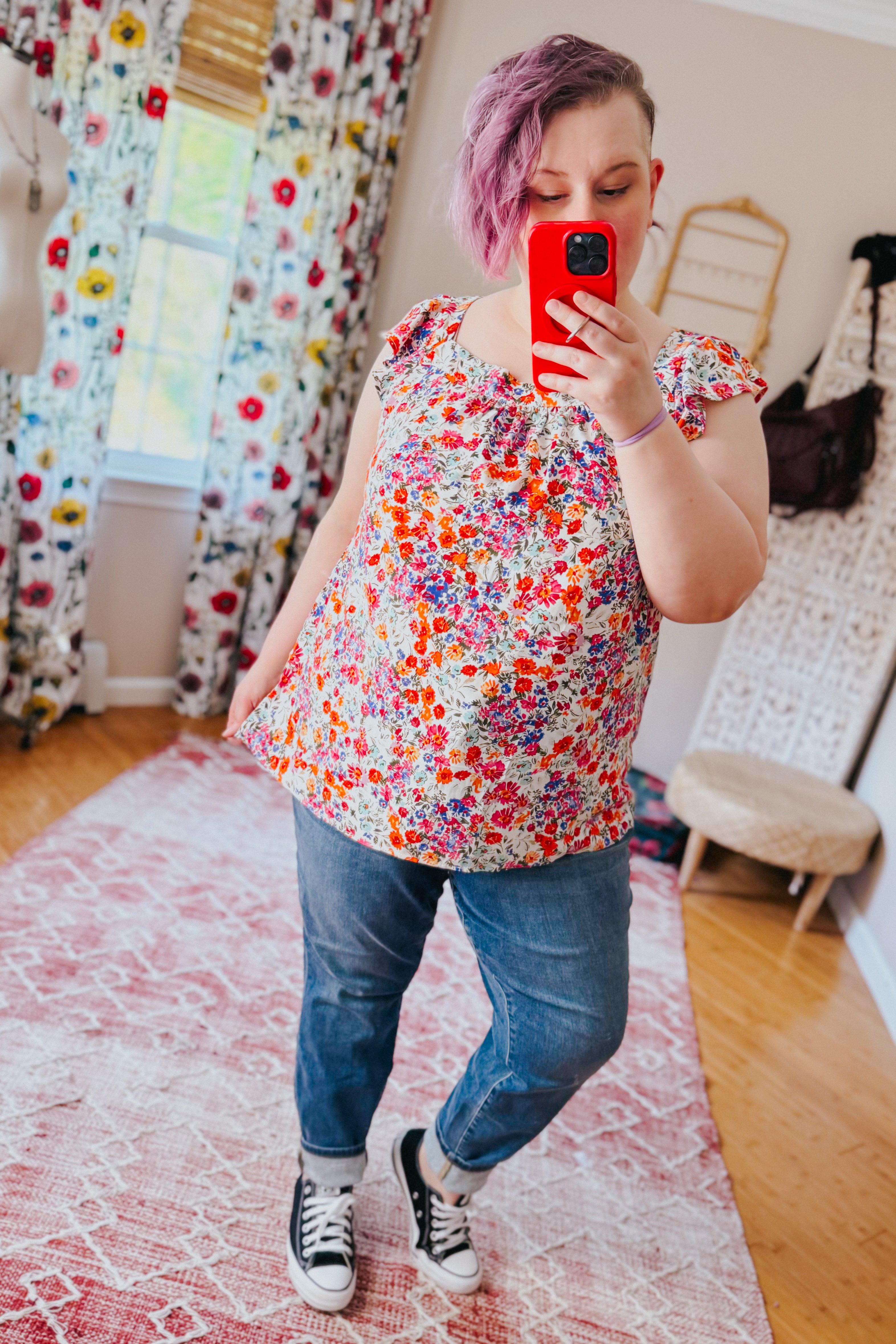 Woman with short pink hair wearing a colorful floral top with ruffle sleeves, jeans, and black sneakers indoors