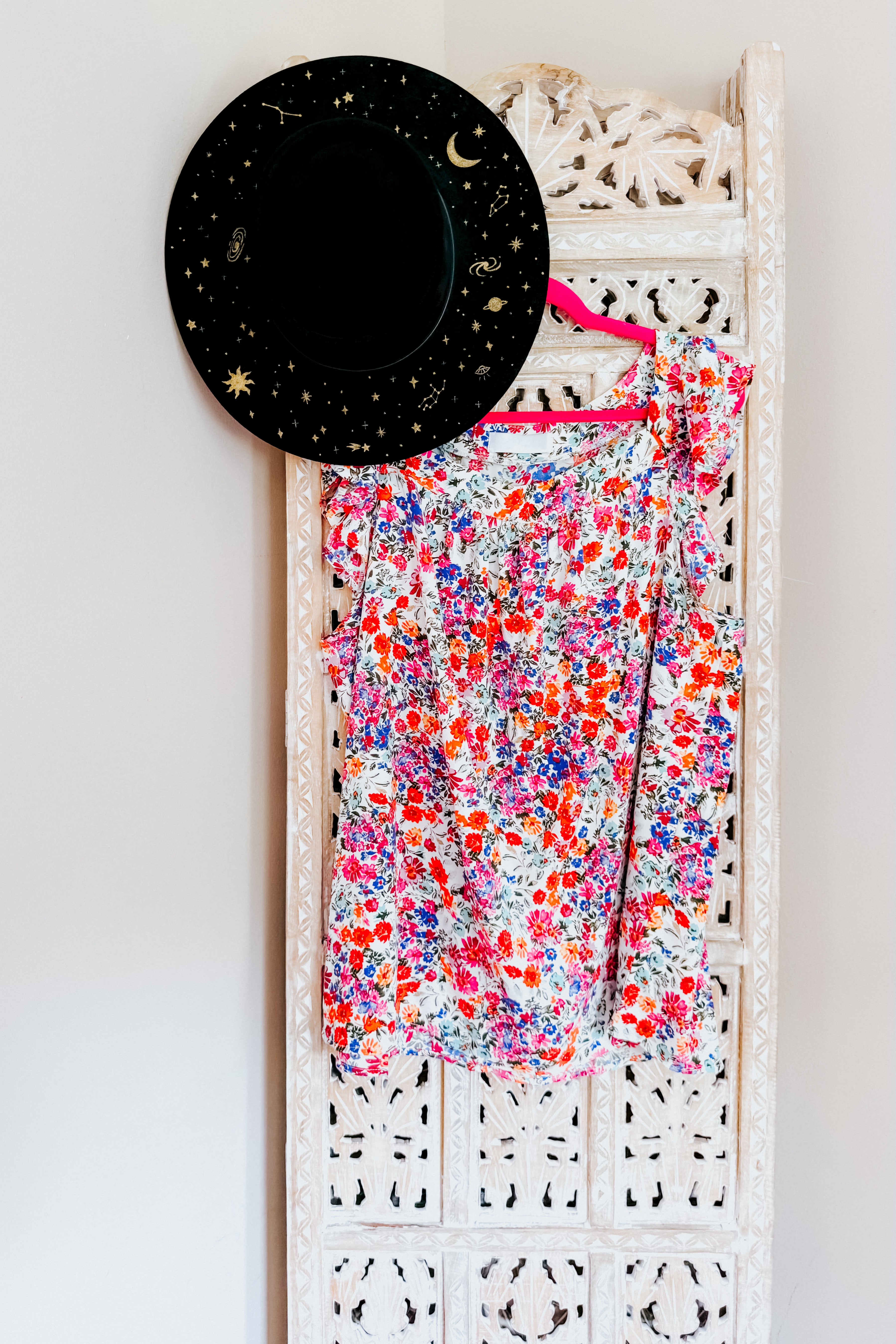 Red floral square neck top hanging on decorative white wood panel beside black hat with gold celestial designs