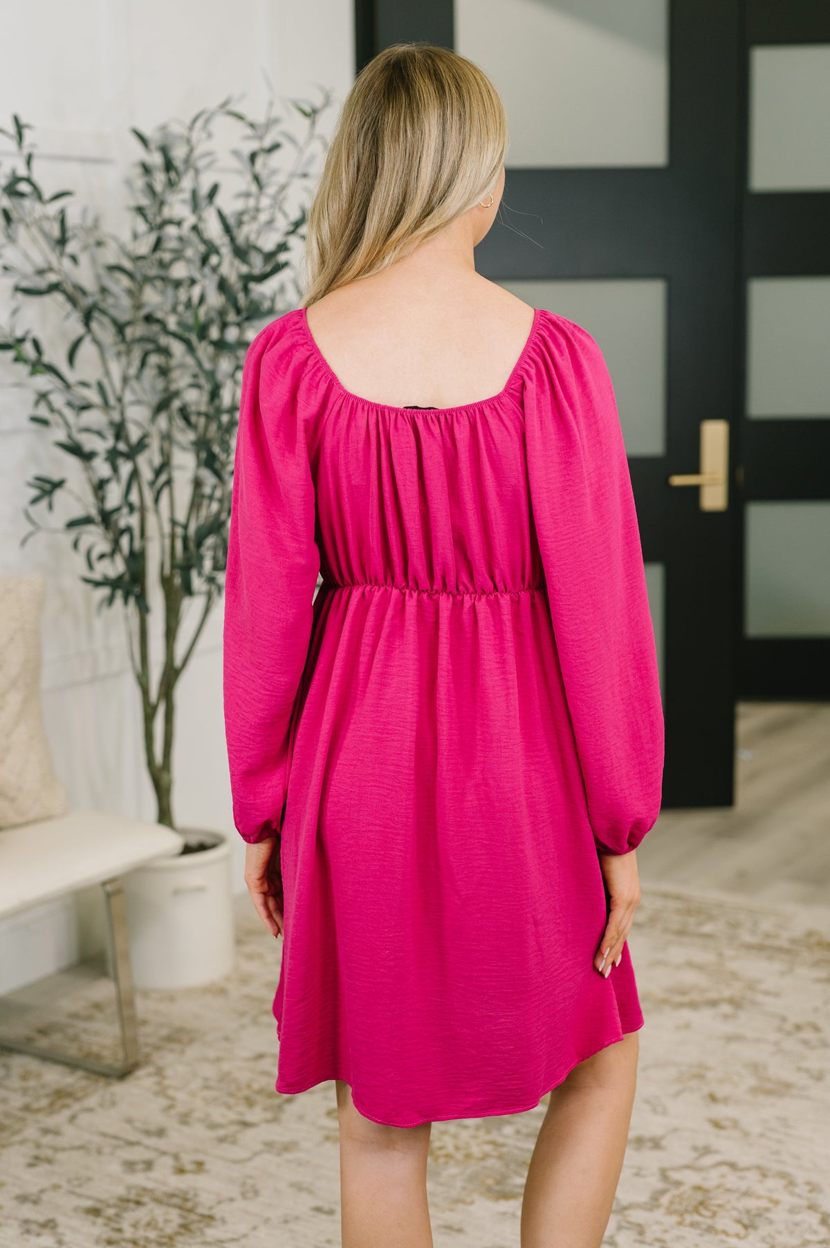 Back view of a woman wearing a pink square neck dress with long raglan sleeves and an elastic waist in a bright home interior