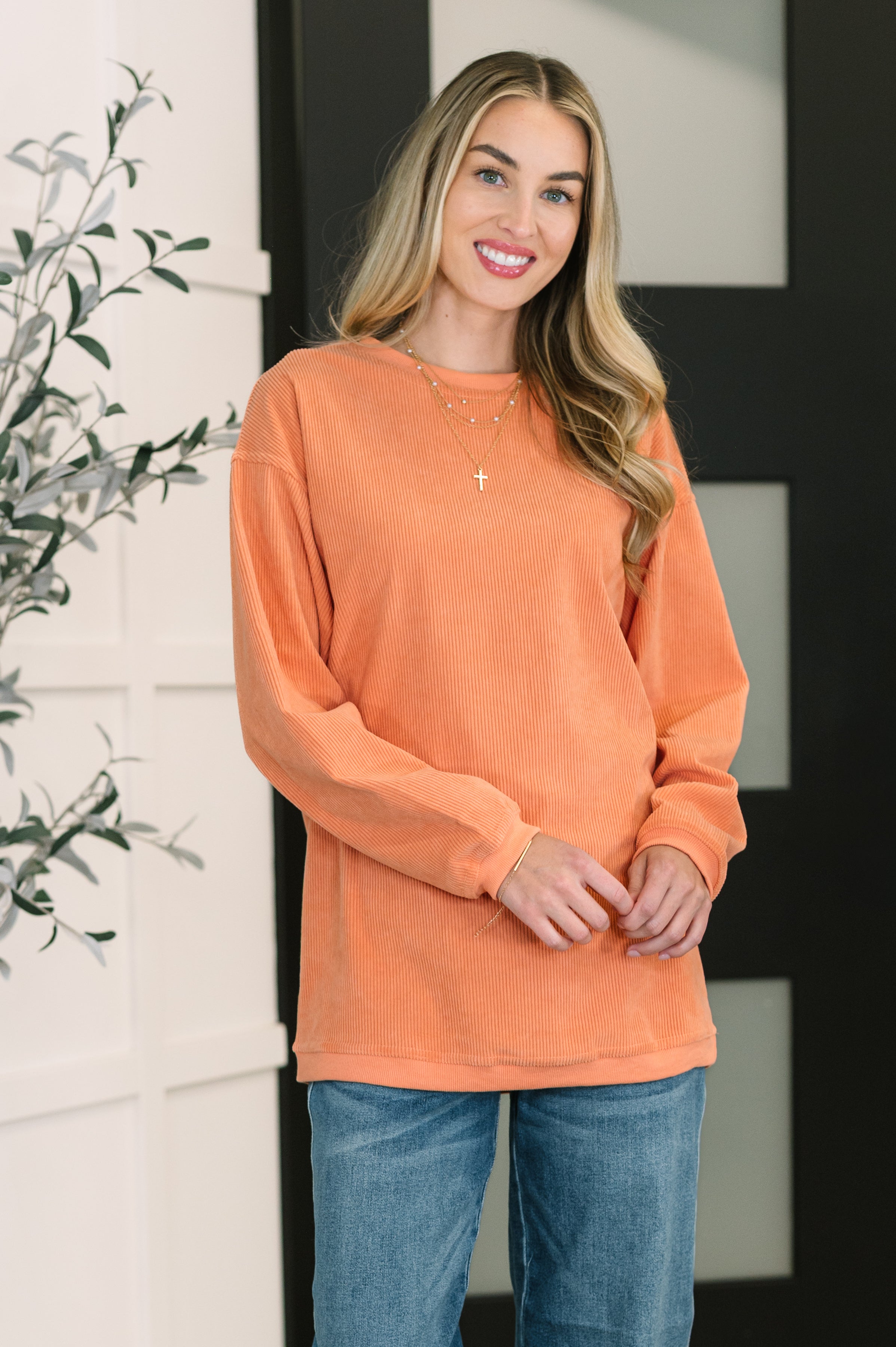 Smiling woman wearing an oversized orange corded sweatshirt paired with blue jeans in a modern indoor setting