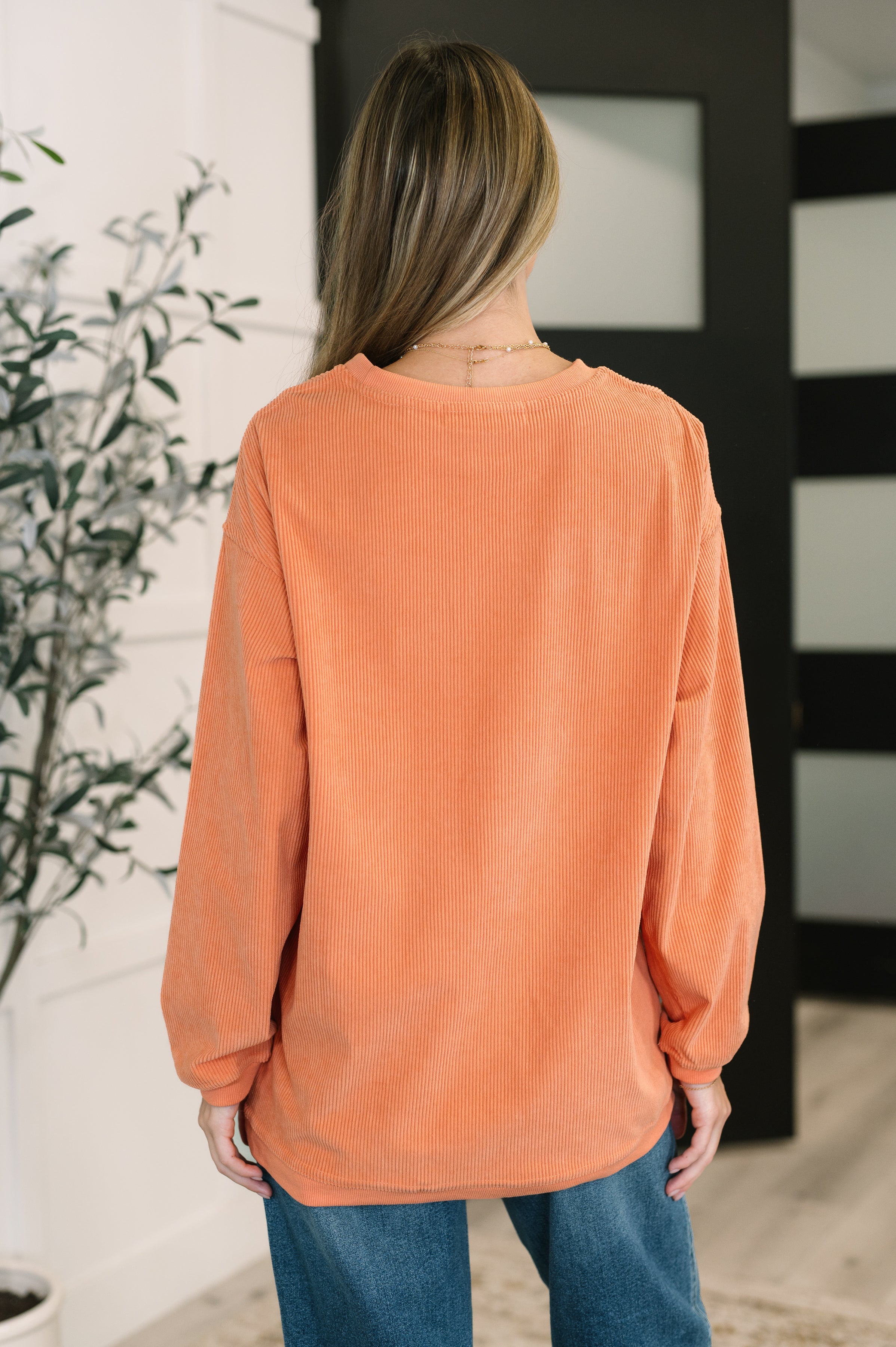 Woman wearing an oversized orange corded sweatshirt with relaxed sleeves, standing indoors near a potted plant and modern door