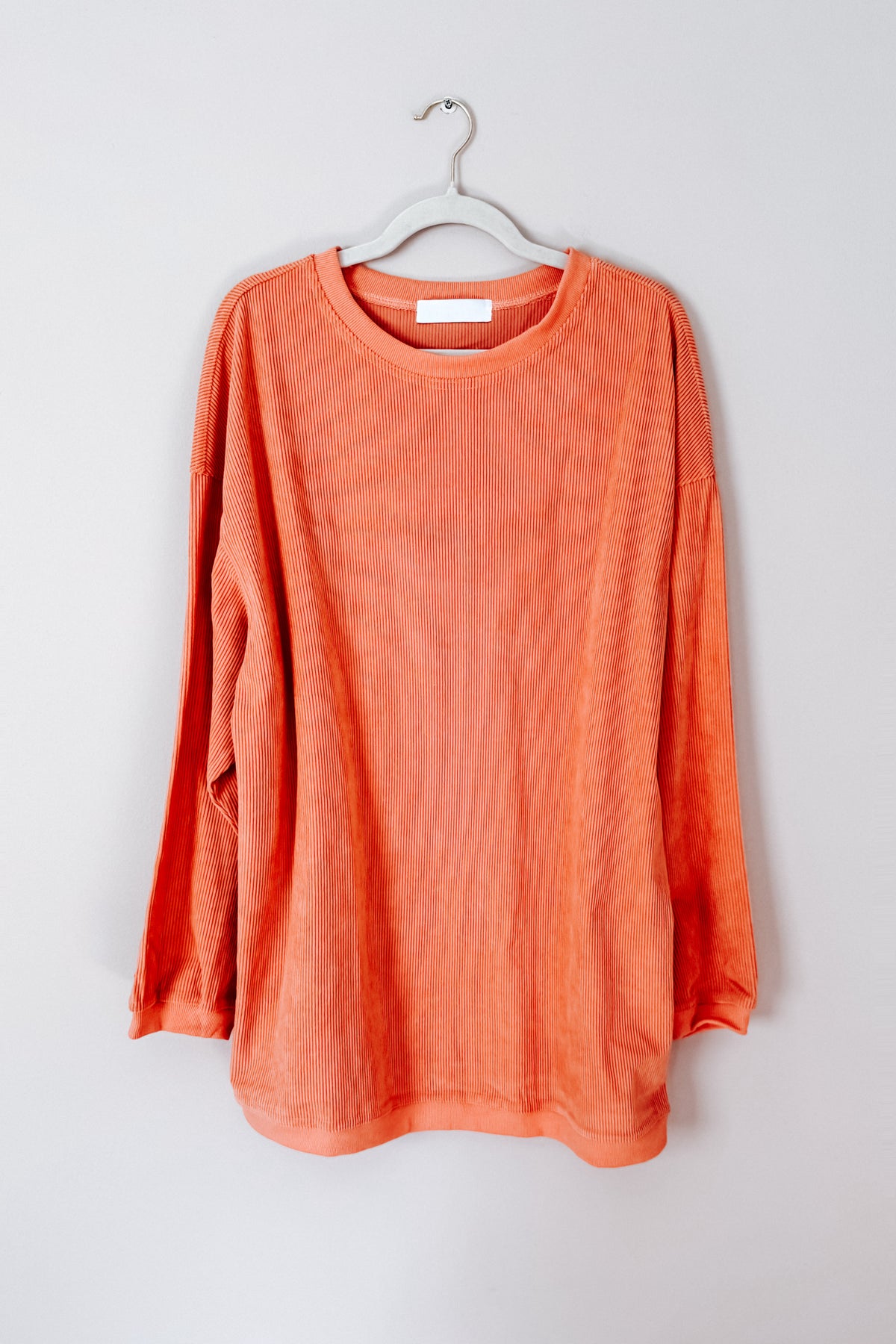 Orange oversized corded sweatshirt with a ribbed round neckline and long sleeves hanging from a metal hanger against a light wall